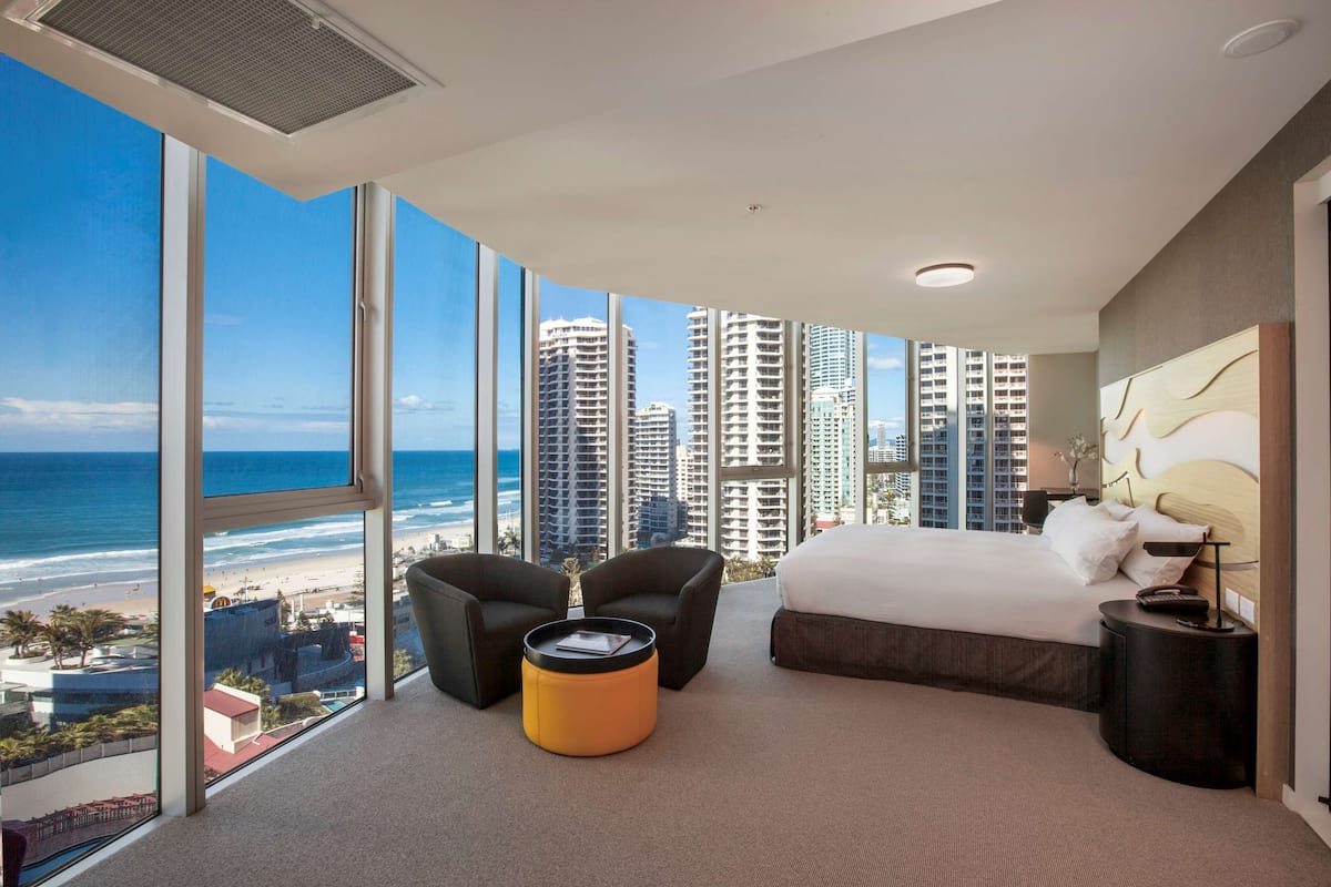 A modern hotel room with a large bed, two armchairs, a small orange ottoman, and a nightstand. The room features large windows offering a view of the beach and high-rise buildings.