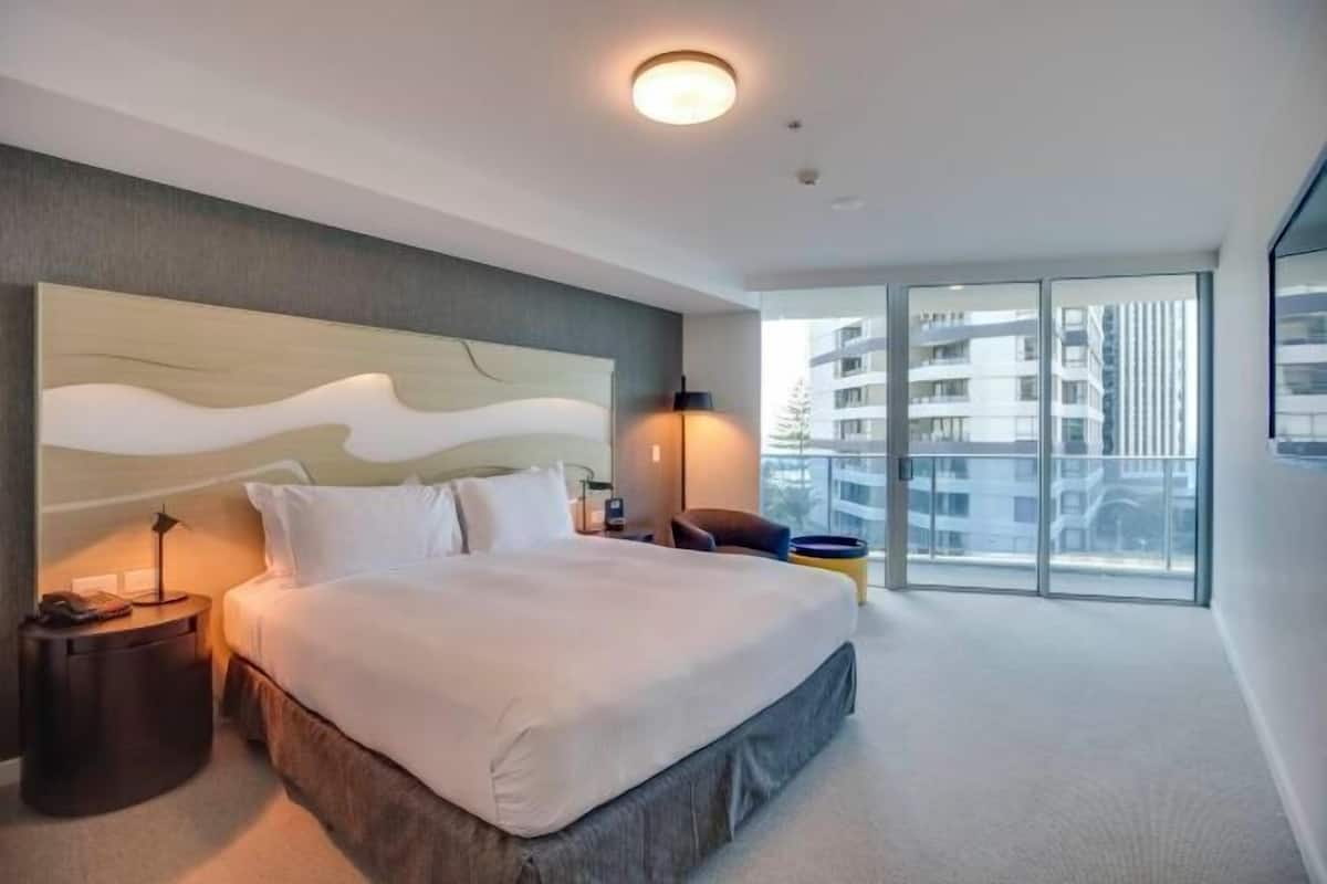 A modern hotel room with a large bed, a nightstand, a lamp, and a view of the city through large windows.