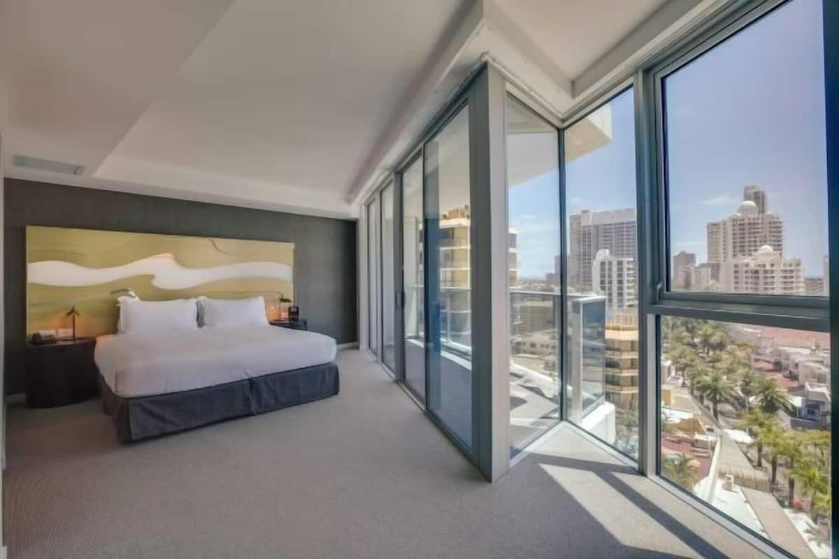 Une chambre d’hôtel moderne dotée d’un grand lit, offrant une vue sur la ville grâce à de larges fenêtres du sol au plafond, et agrémentée d’une tête de lit contemporaine.