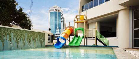 Una piscina con tobogán y estructura de juegos, rodeada por un edificio de diseño moderno y vista a un rascacielos alto al fondo.