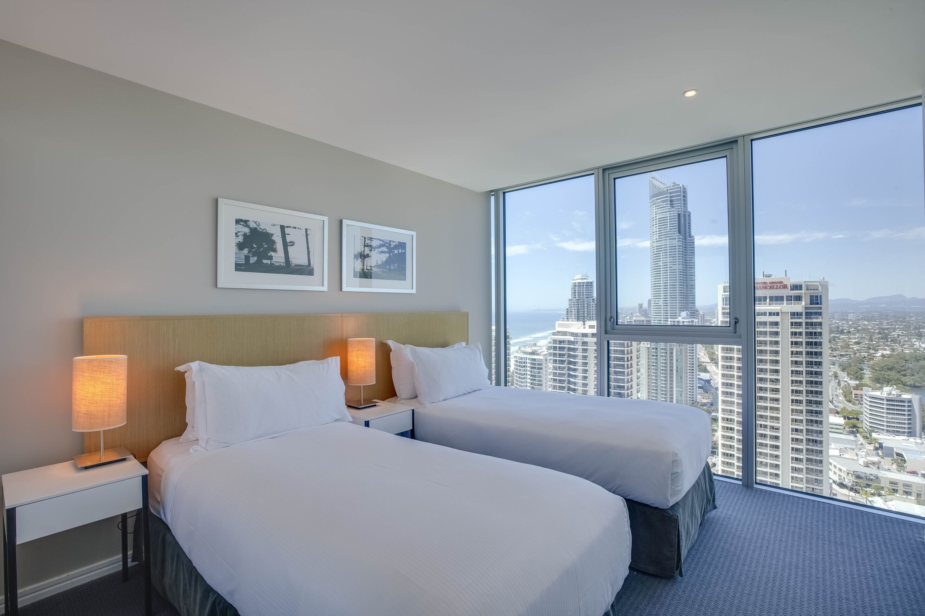 A hotel room with two beds, a nightstand with a lamp, and a large window offering a city view.