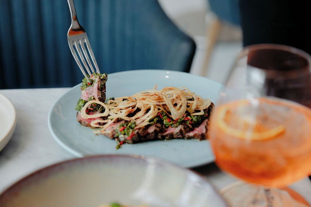 A plate with a slice of meat topped with onions and greens, a fork, and a glass of orange drink.