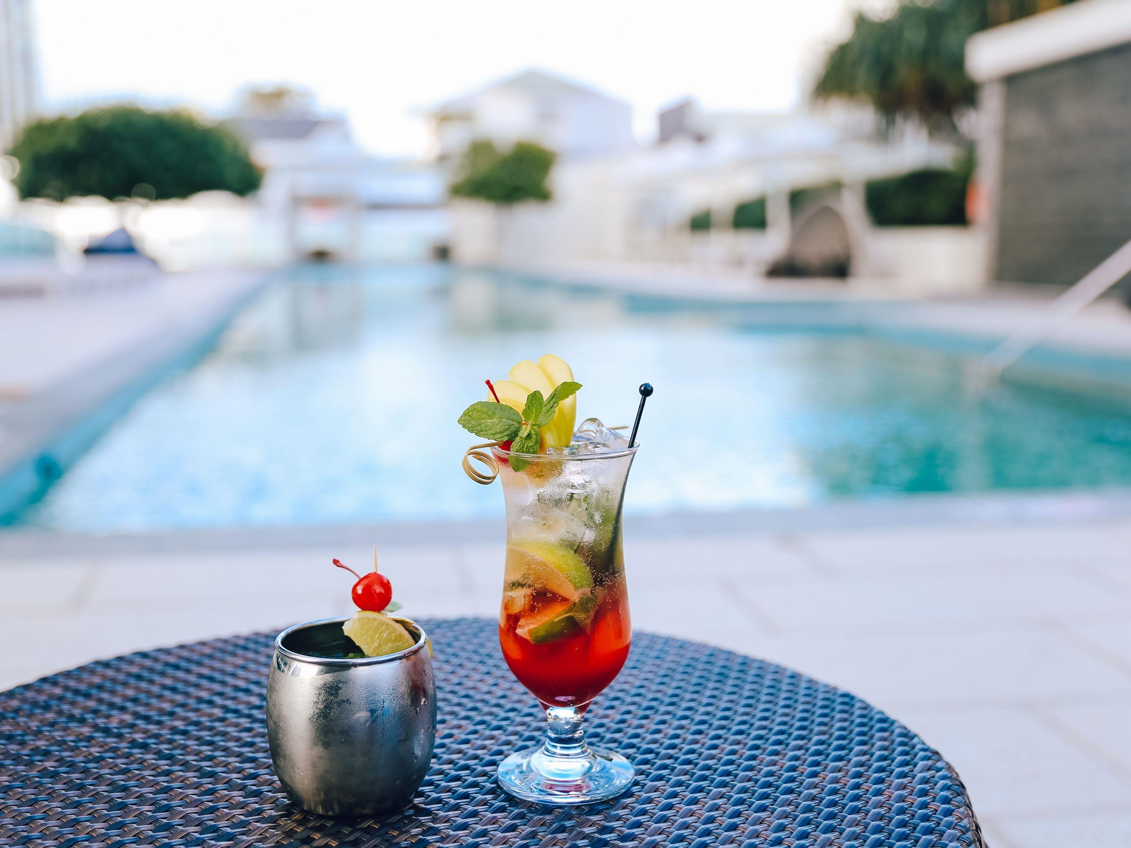 A cocktail on a table by a poolside with a clear view of the pool and buildings in the background.