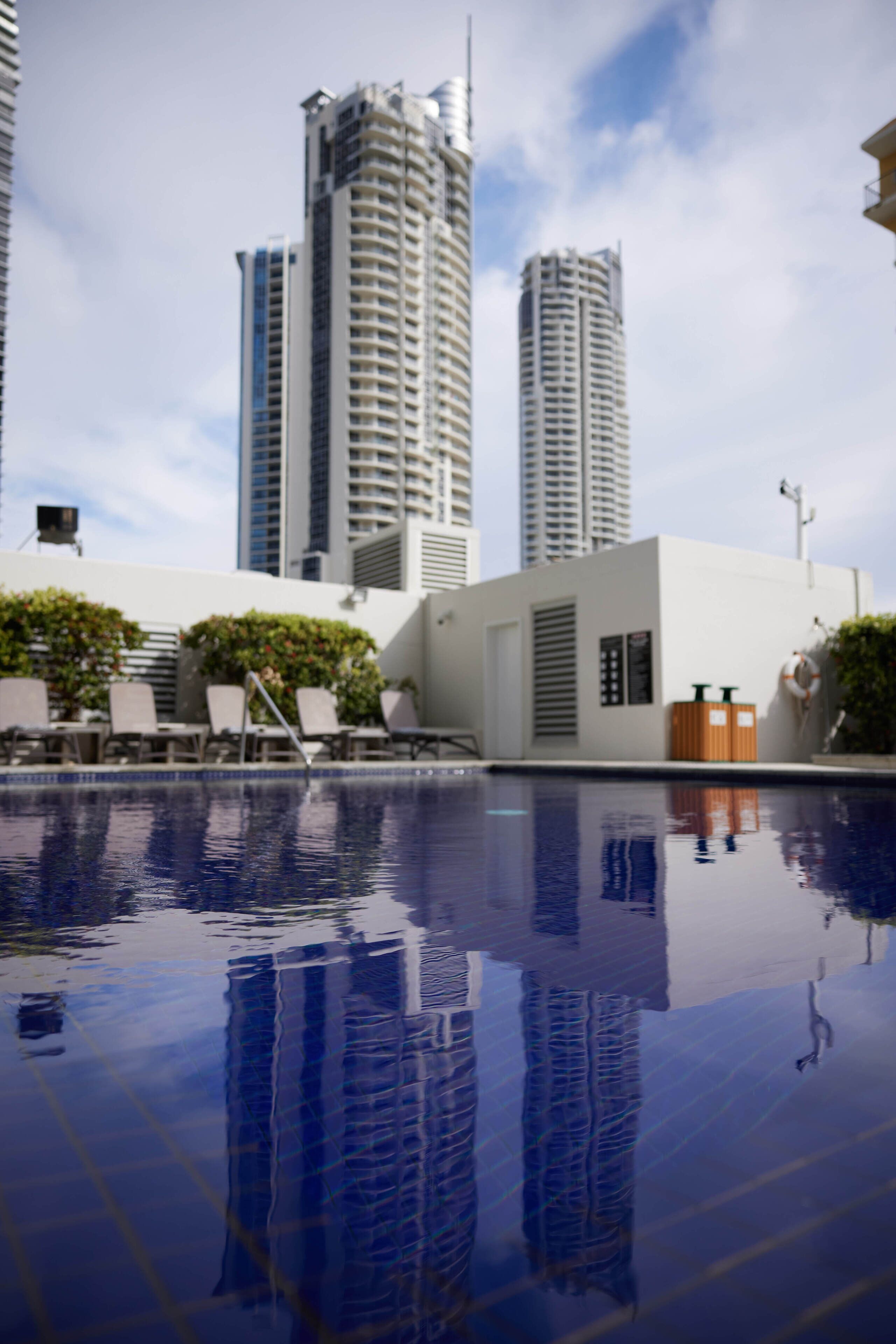 A swimming pool with reflections of tall buildings, lounge chairs, and a small structure with a door and windows.