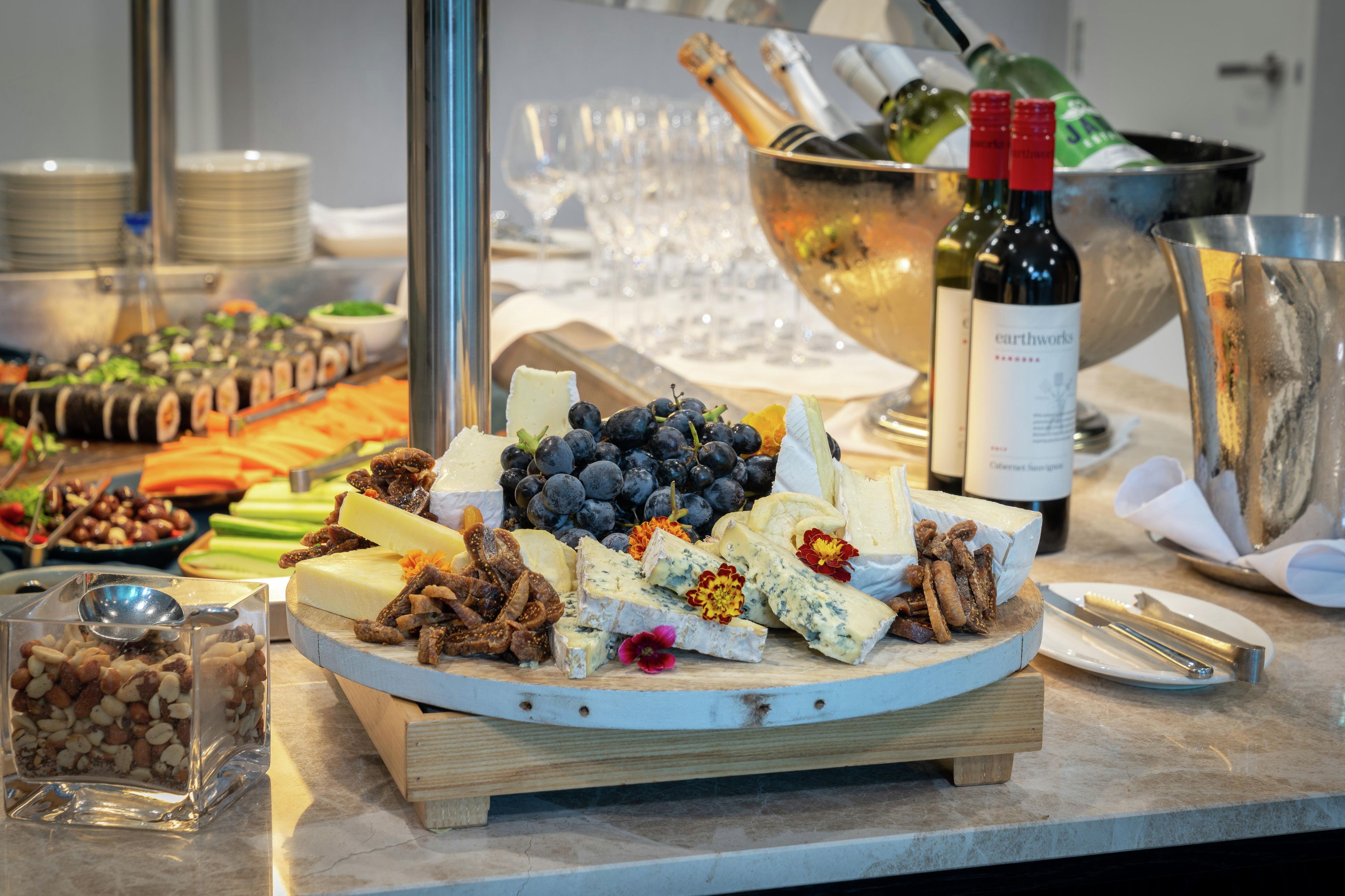 A cheese and fruit platter on a wooden board, with a bottle of wine and a bowl of champagne in the background.