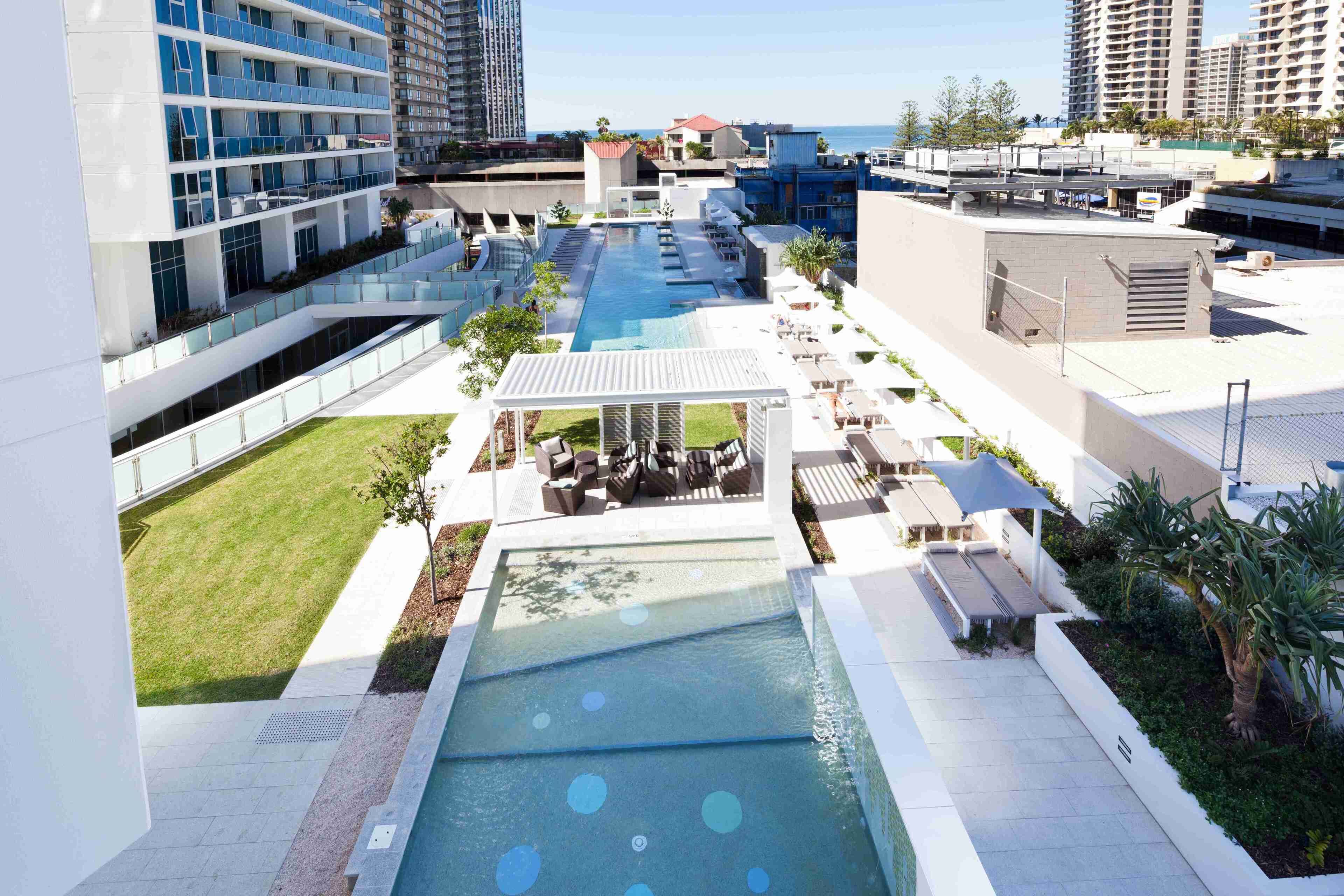 A rooftop pool area with lounge chairs and umbrellas, surrounded by modern buildings and a view of the ocean.