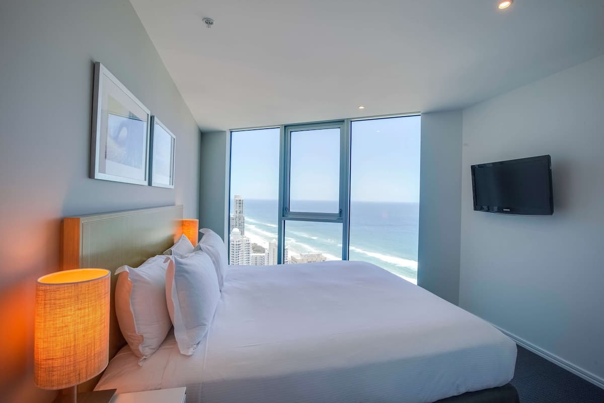 A hotel room with a large bed, a TV mounted on the wall, and a view of the ocean through large windows.