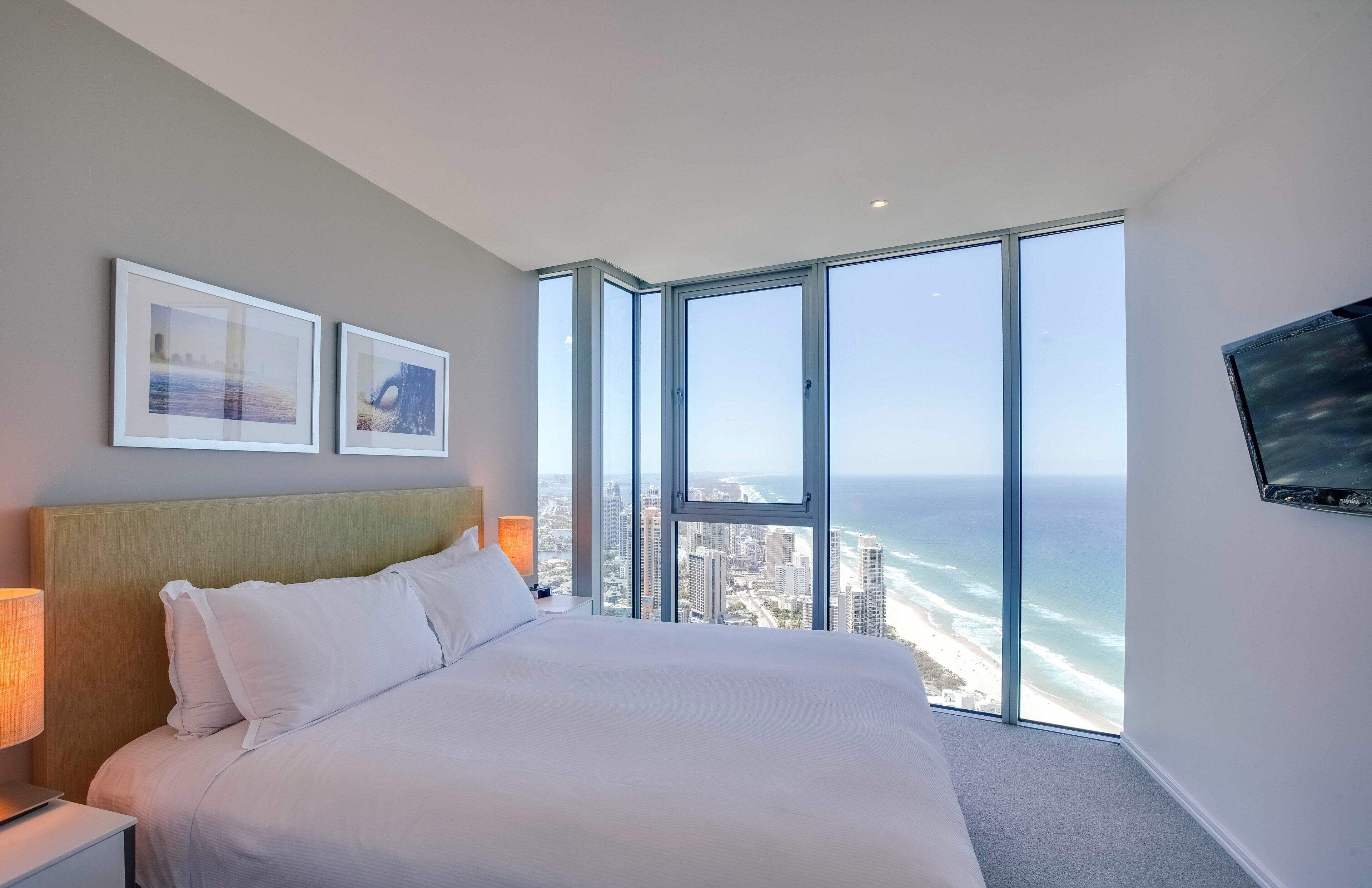 Quarto de hotel com uma cama grande, duas fotos emolduradas na parede, uma TV montada e vista para o oceano e o horizonte da cidade através de grandes janelas.