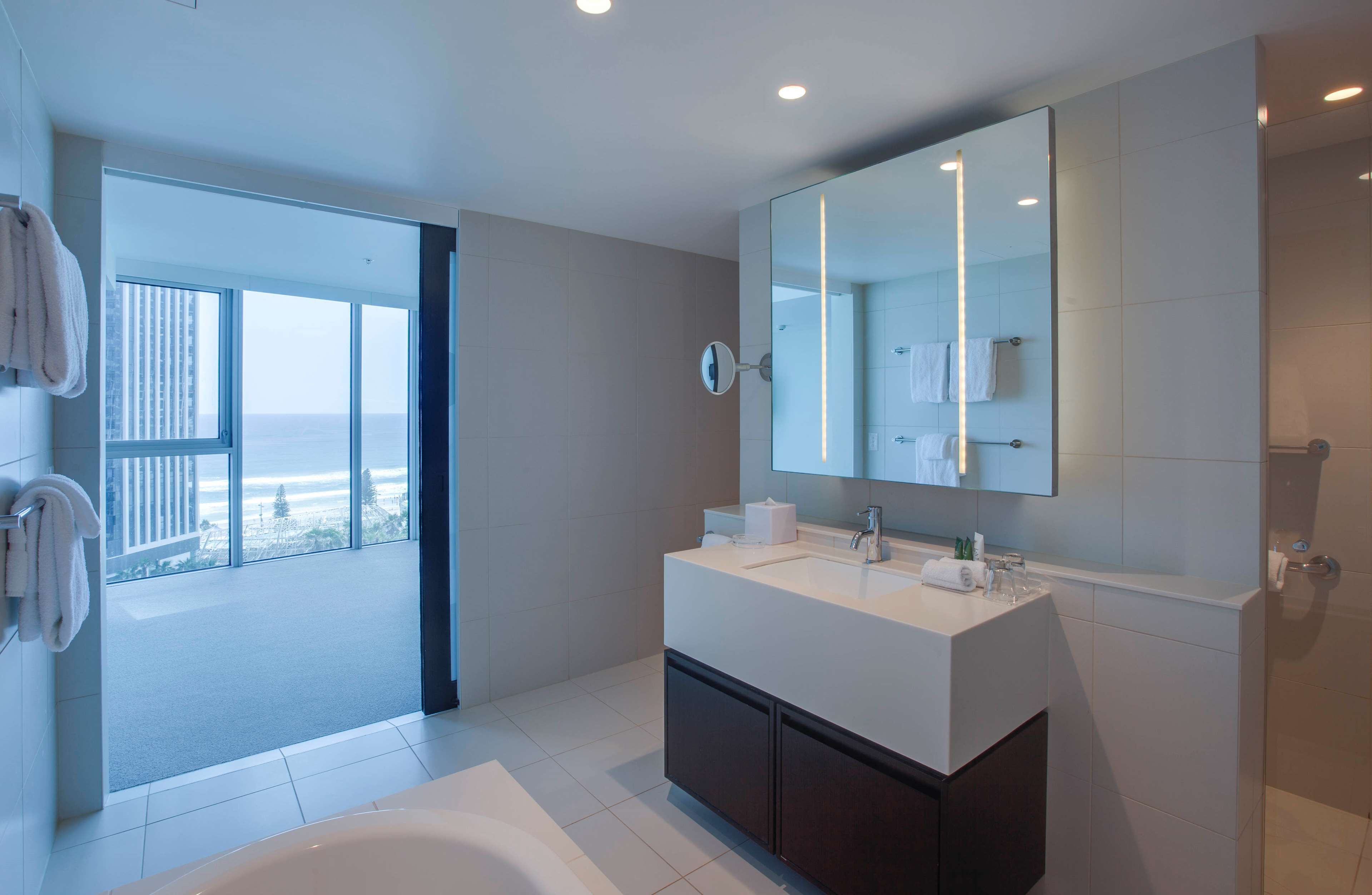 A modern bathroom with a large mirror, a bathtub, and a sink. There is a view of the beach through the glass door.
