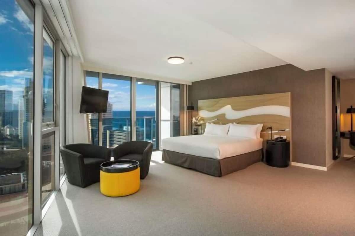 A modern hotel room with a large bed, a flat-screen TV, a sofa, and a yellow ottoman.