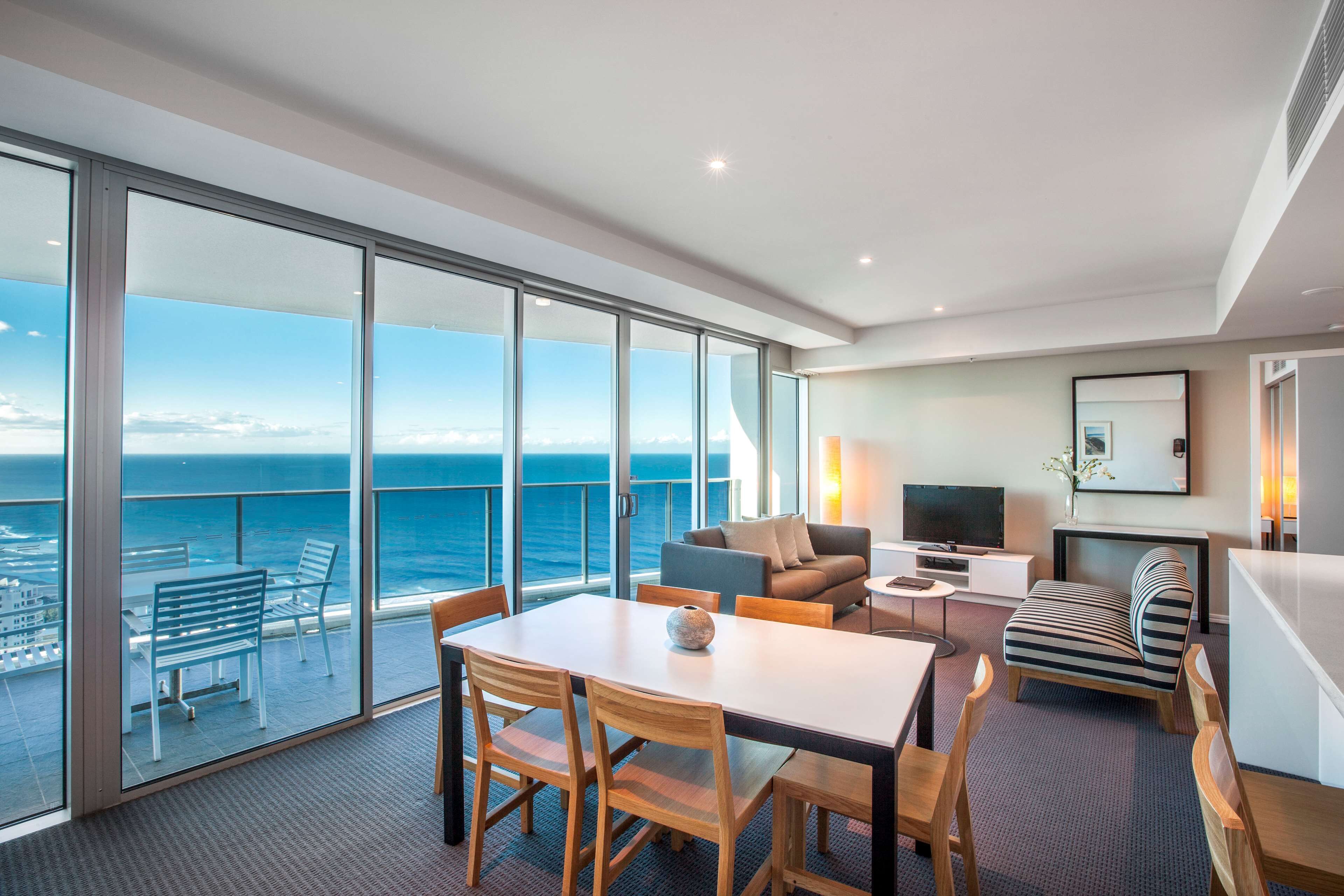 Una habitación moderna con un gran ventanal con vistas al mar, una zona de comedor con sillas de madera, un sofá y un televisor sobre un mueble.