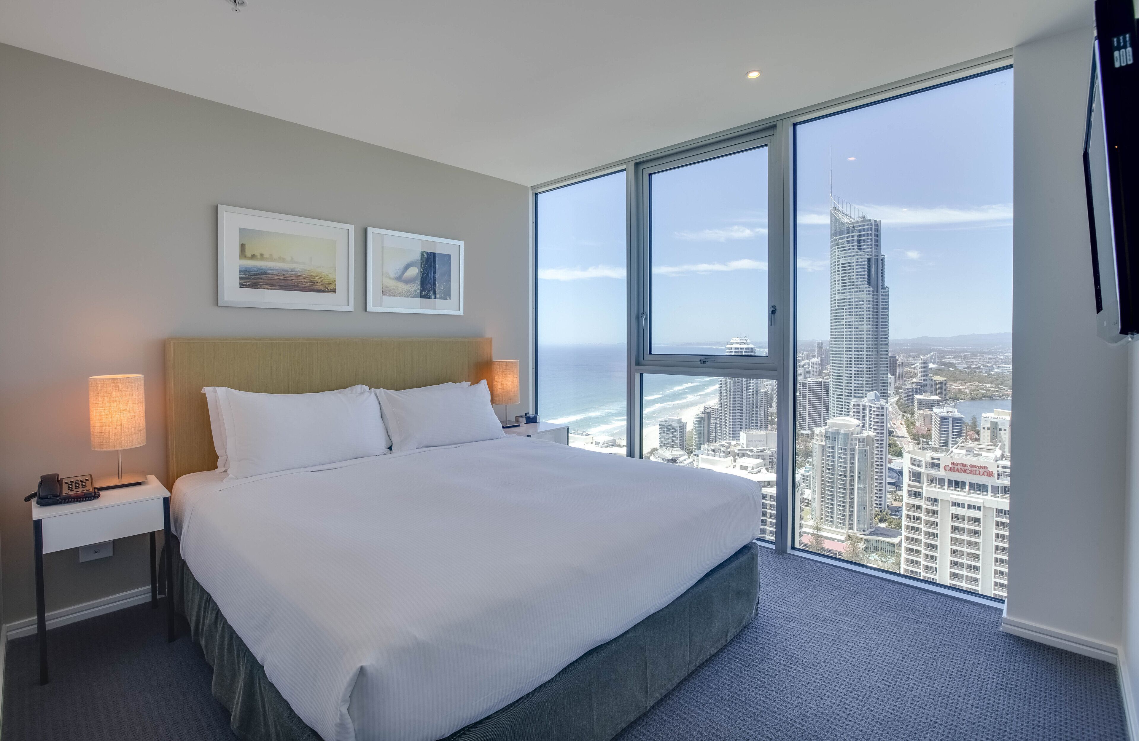 A hotel room with a large bed, two bedside tables with lamps, and a view of a cityscape through floor-to-ceiling windows.
