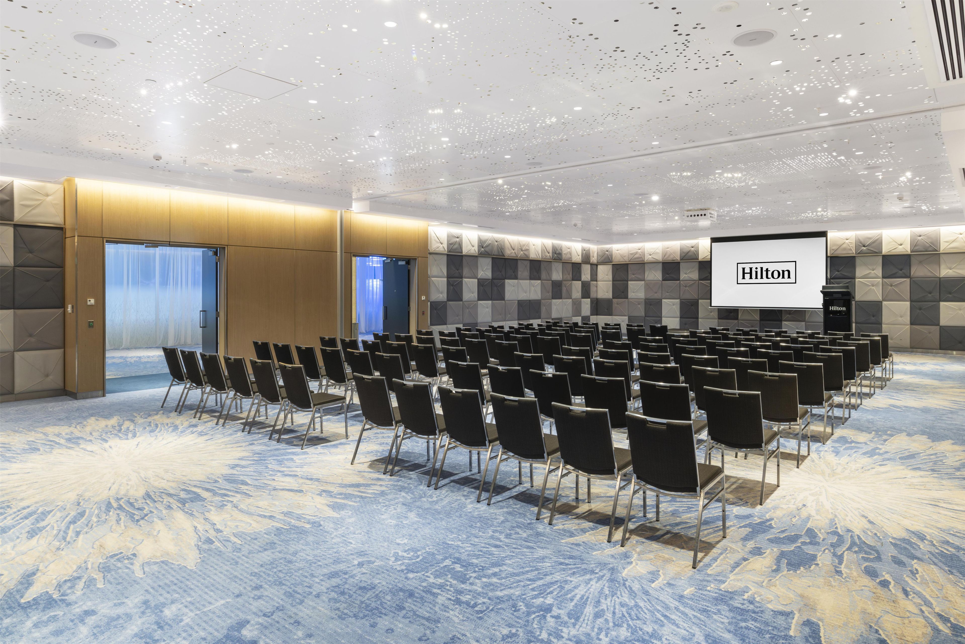 A conference room with rows of chairs, a podium, and a screen displaying 'Hilton' branding.
