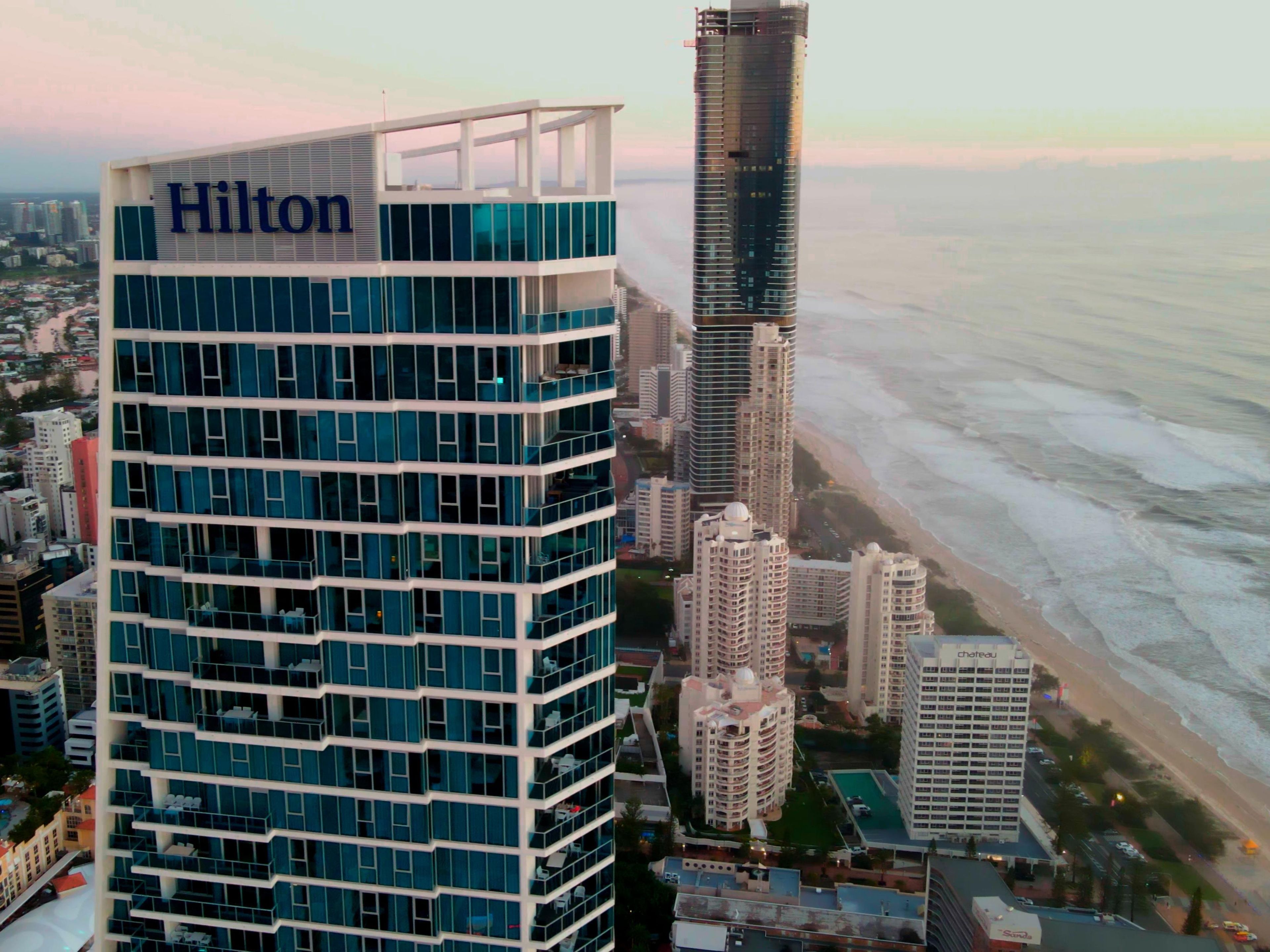 Um hotel alto com um grande letreiro 'Hilton', rodeado por outros edifícios e uma praia ao fundo.