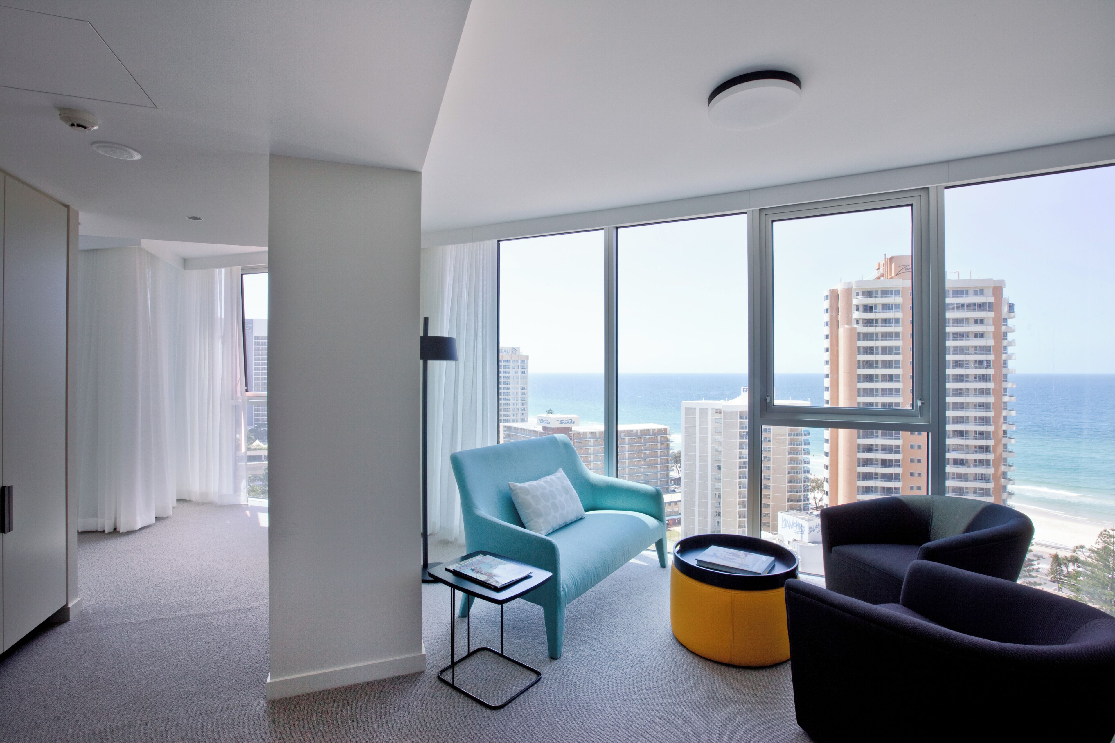 A modern hotel room with a large window, a teal armchair, a black armchair, and a small yellow ottoman.