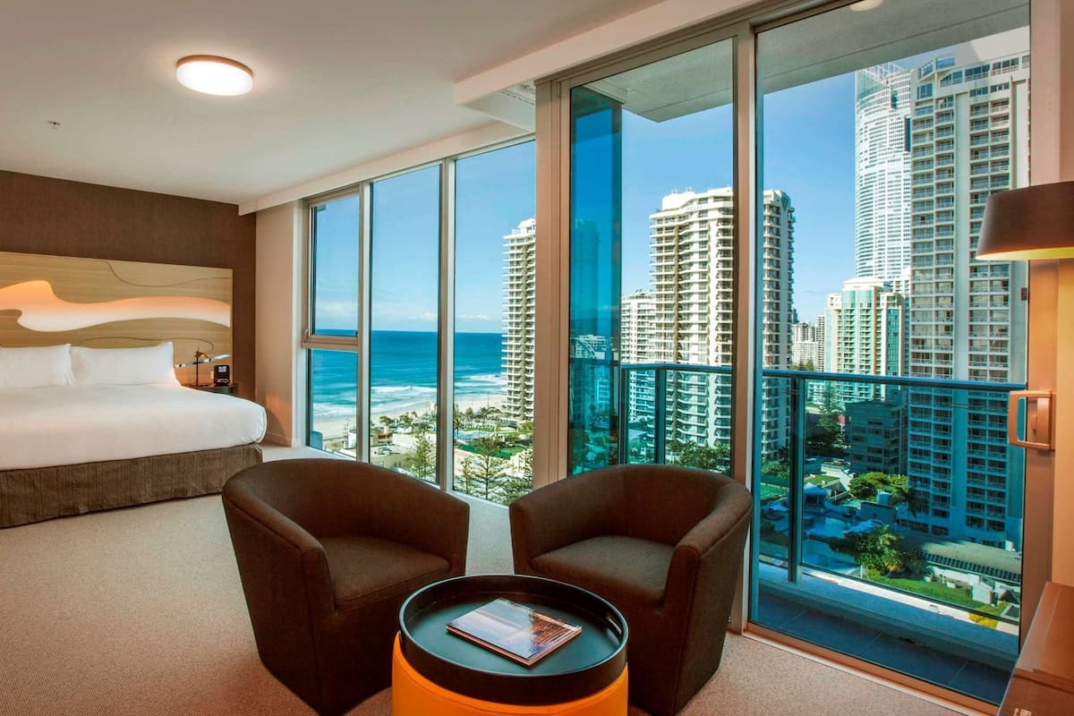 A hotel room with a large bed, two armchairs, a small table, and a view of tall buildings and the ocean.