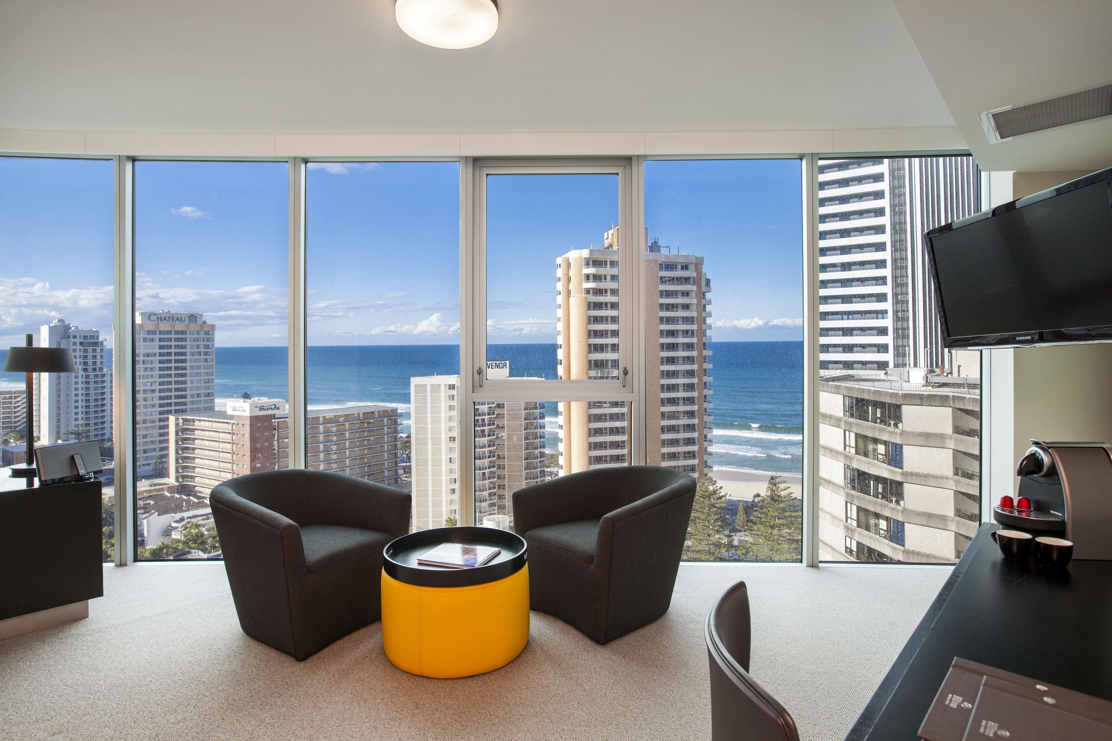 A modern hotel room with a large window offering a view of the beach and ocean, two armchairs, a yellow ottoman, a flat-screen TV, and a coffee machine.