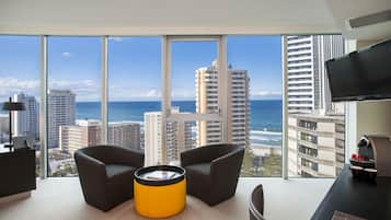 A modern hotel room with a large window offering a view of the beach and ocean, two armchairs, a yellow ottoman, a flat-screen TV, and a coffee machine.