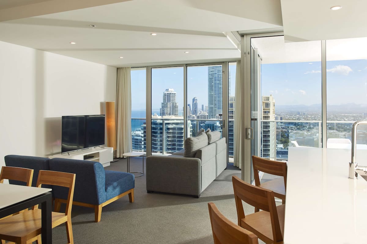 A modern hotel room with a large balcony, a flat-screen TV, a sofa, a dining table, and a city view.
