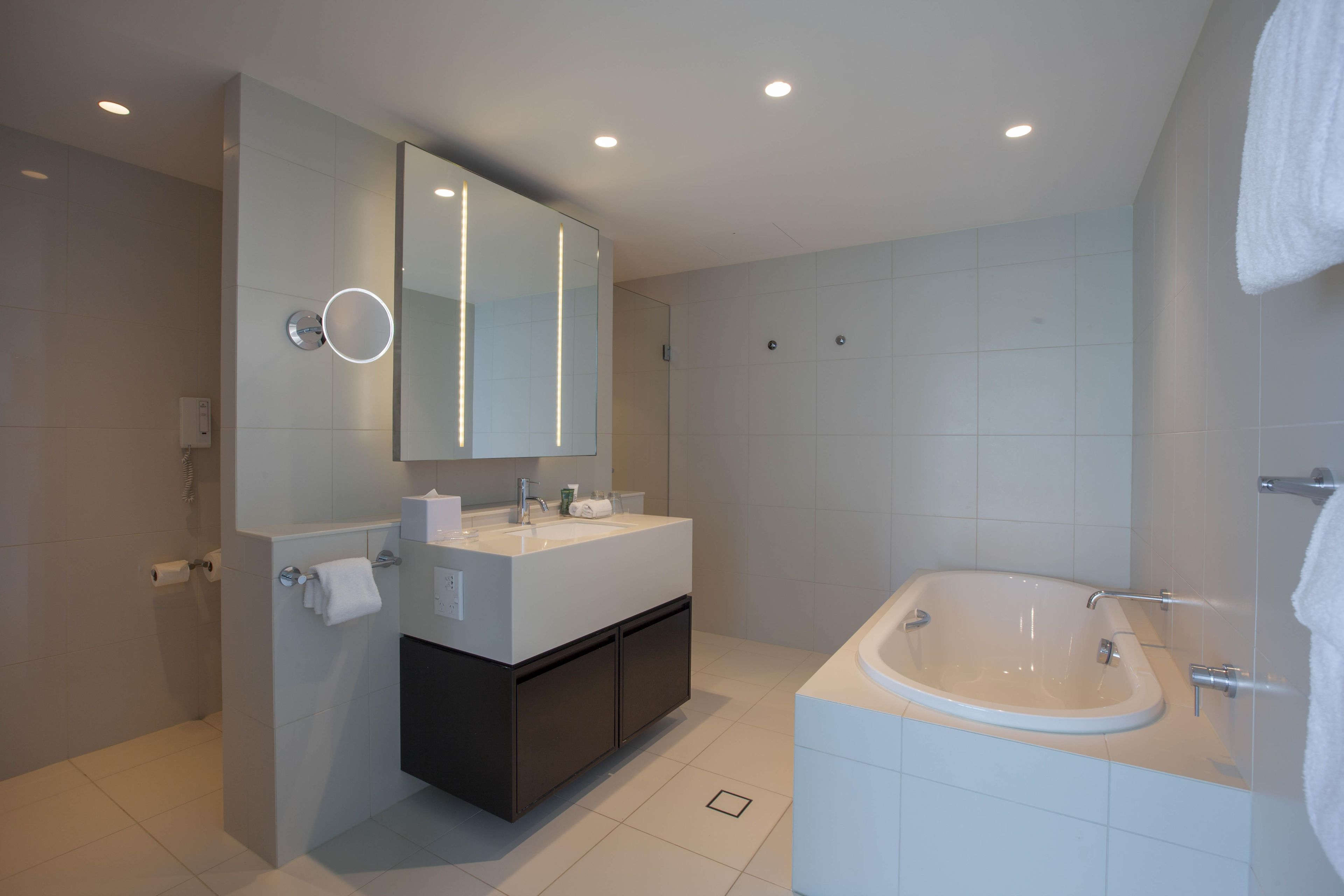 A modern bathroom with a large mirror, a double sink vanity, a bathtub, and recessed ceiling lights.