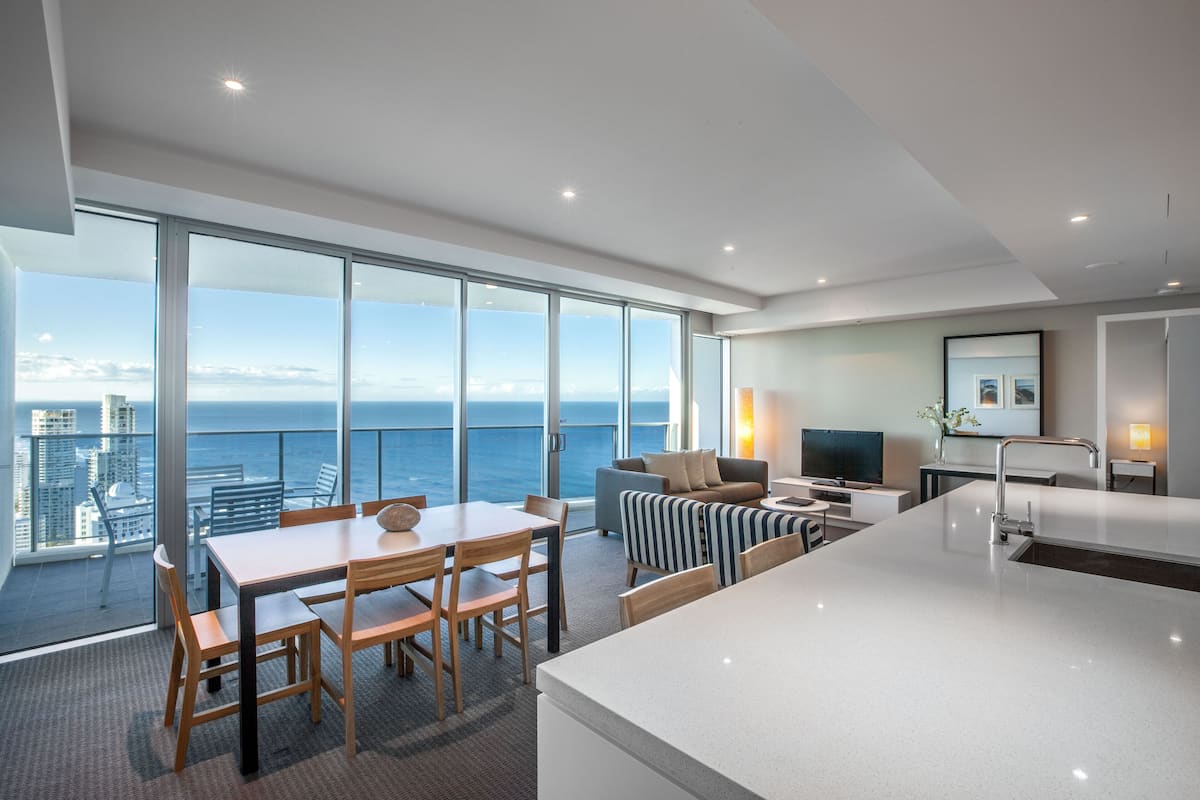A modern hotel room with a large balcony, a dining area, a sofa, a TV, and a kitchen area.