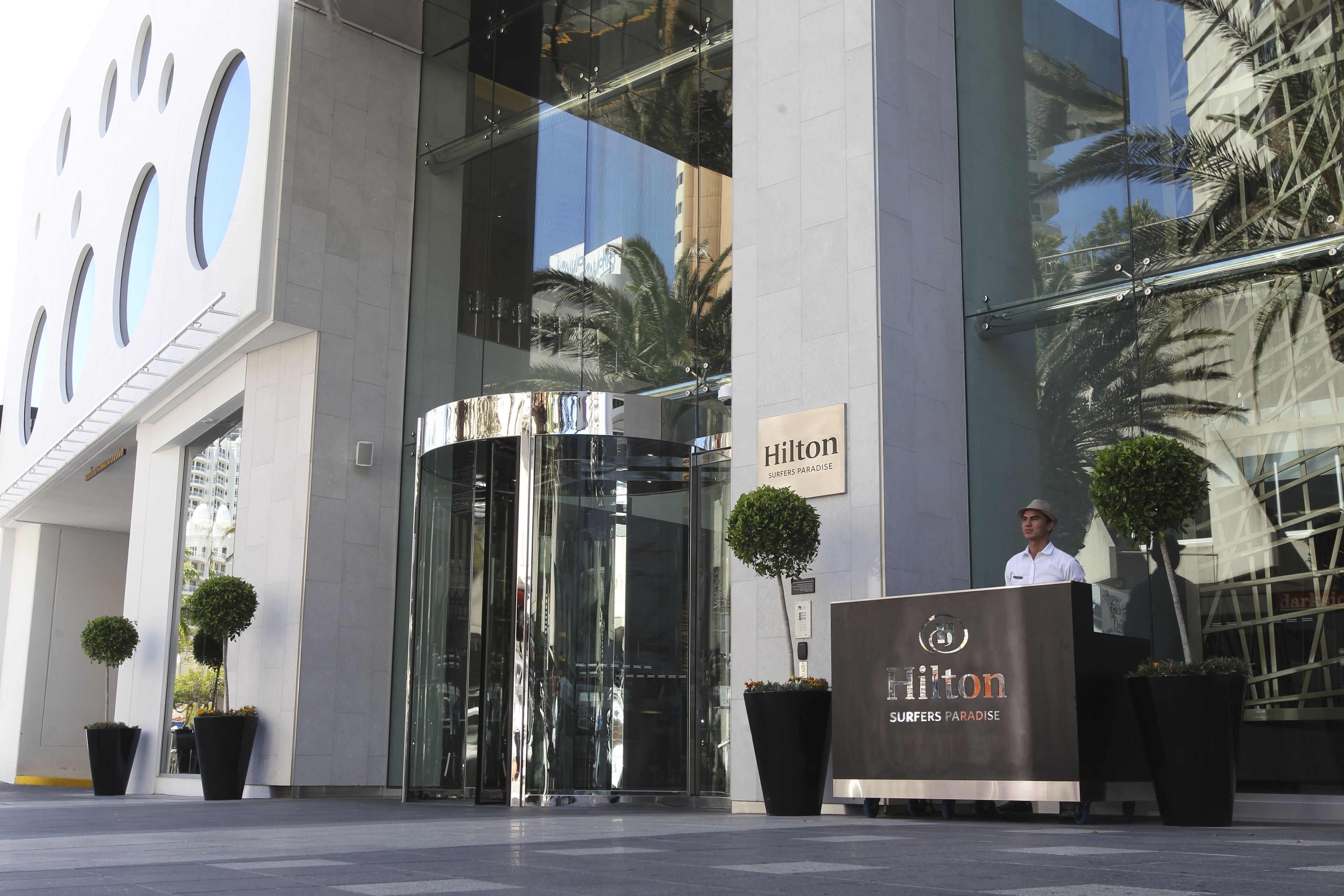 Entrada de hotel com porta giratória de vidro, um pedestal com o nome do hotel e plantas em vasos.
