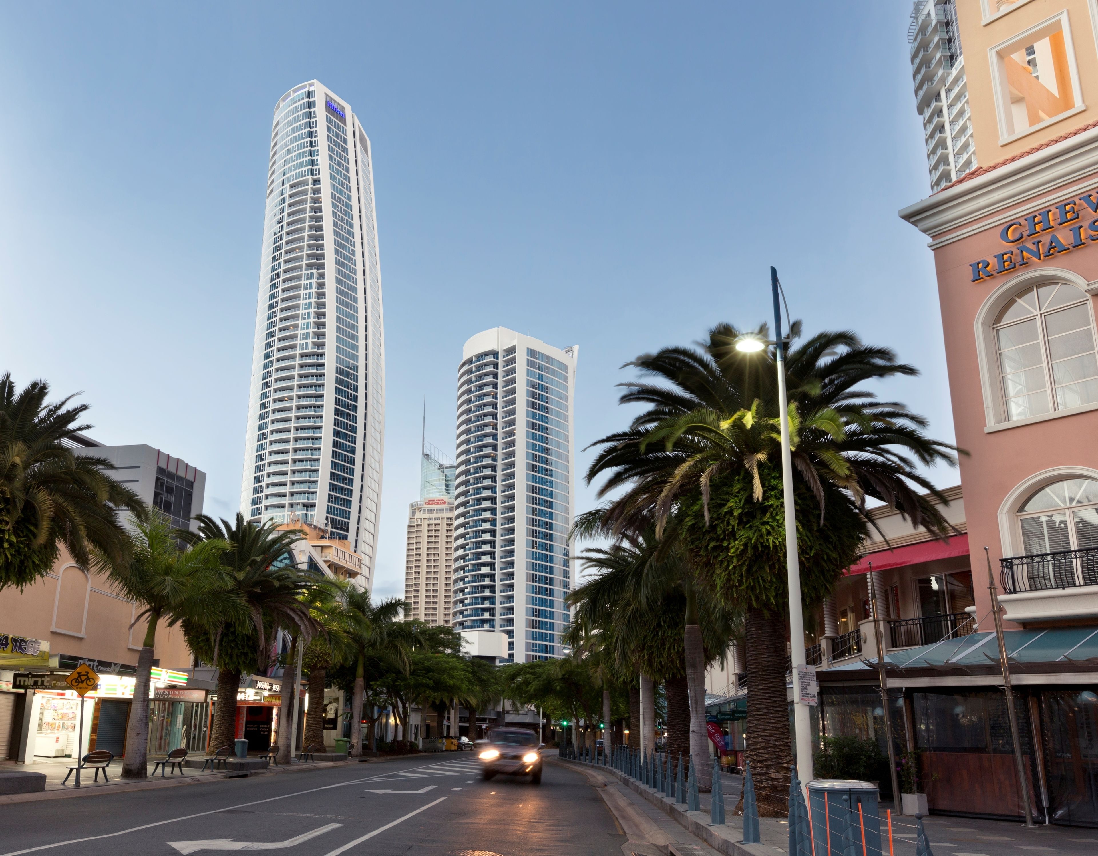 A city street with tall buildings, palm trees, and a car driving.