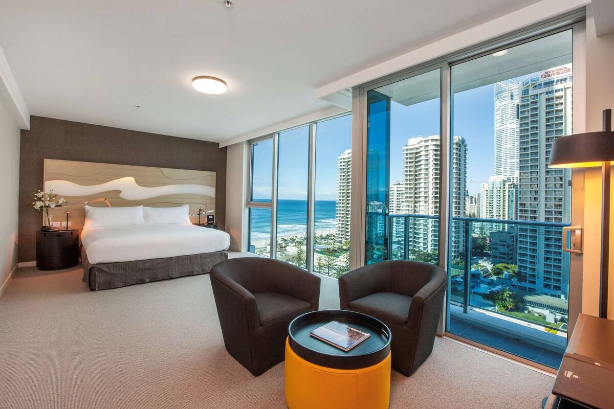 A modern hotel room with a large bed, two armchairs, a small table, and a view of the city and ocean.