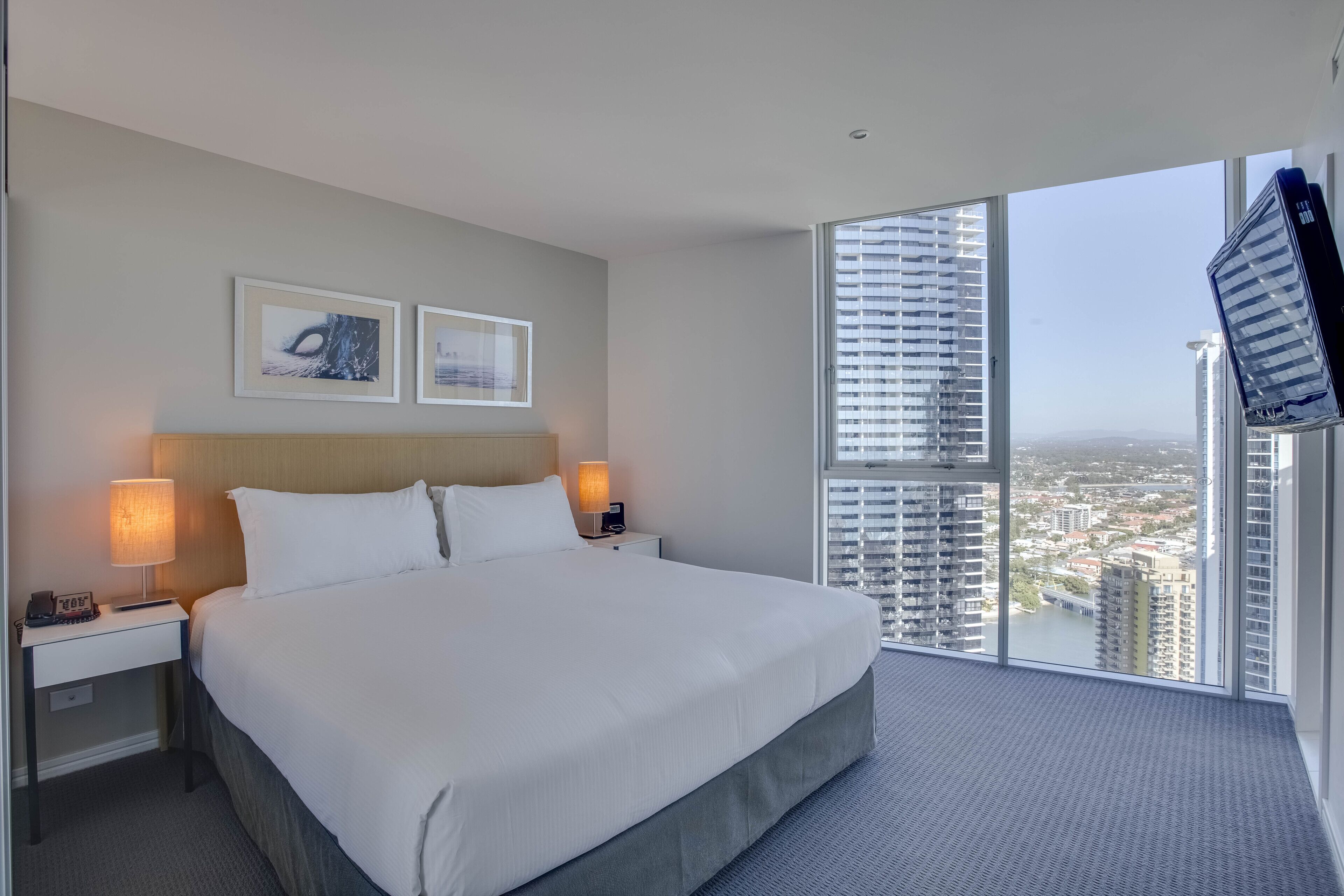 Habitación de hotel con una cama grande, dos mesitas de noche con lámparas, un televisor en la pared y vistas a la ciudad a través de amplios ventanales.