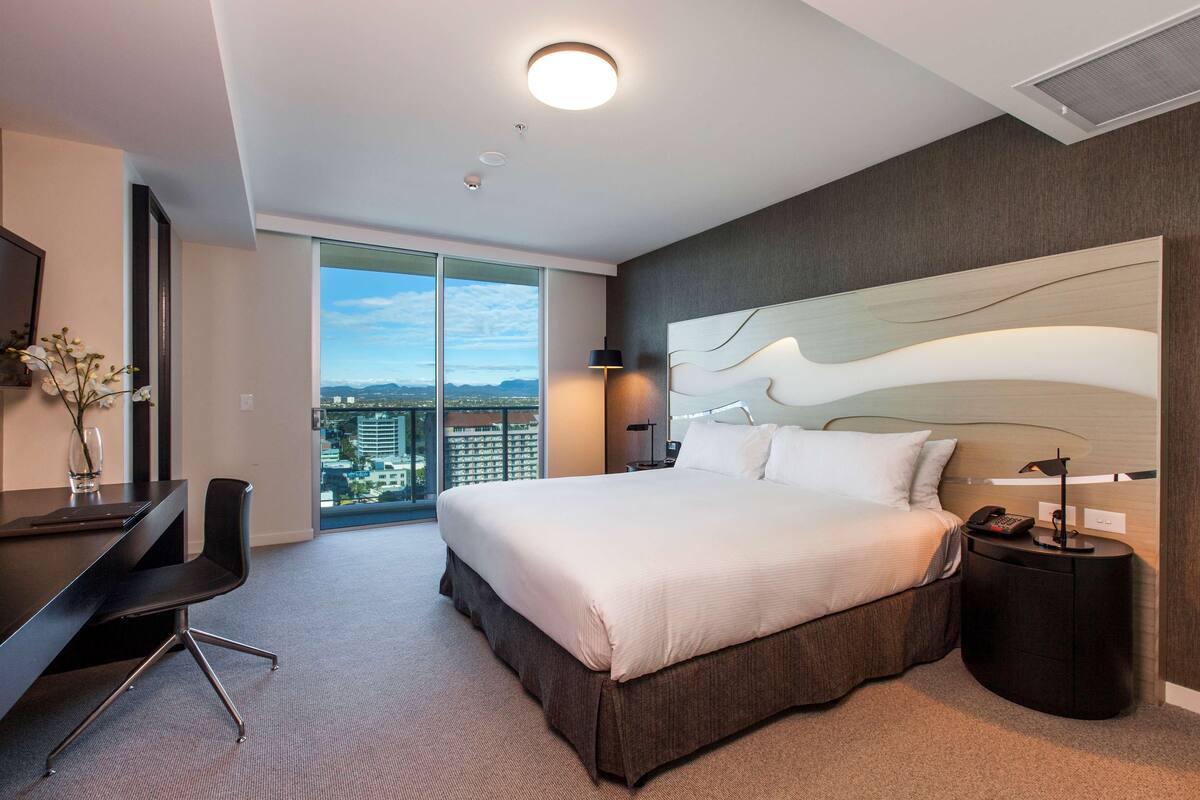 A modern hotel room with a large bed, a desk, a chair, a nightstand, and a view of the city.