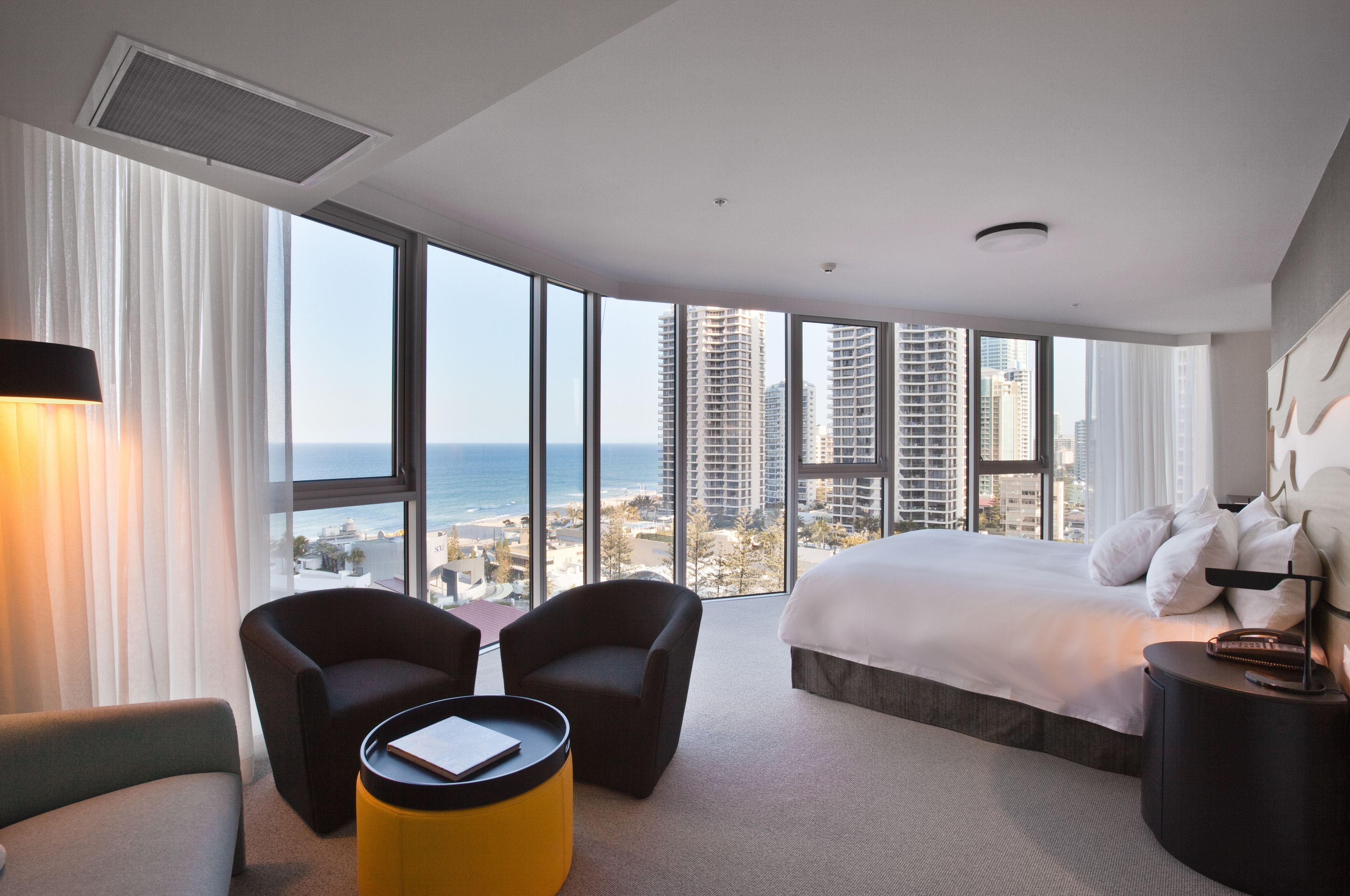 A hotel room with a large bed, two armchairs, a small table, and a view of the city and ocean.