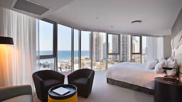 A hotel room with a large bed, two armchairs, a small table, and a view of the city and ocean.