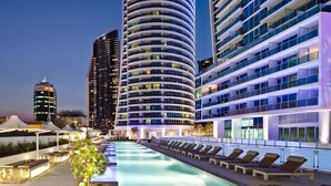 Een modern hotel met een gebogen gevel, een zwembad op het dak met ligbedden, en een stadsgezicht op de achtergrond.