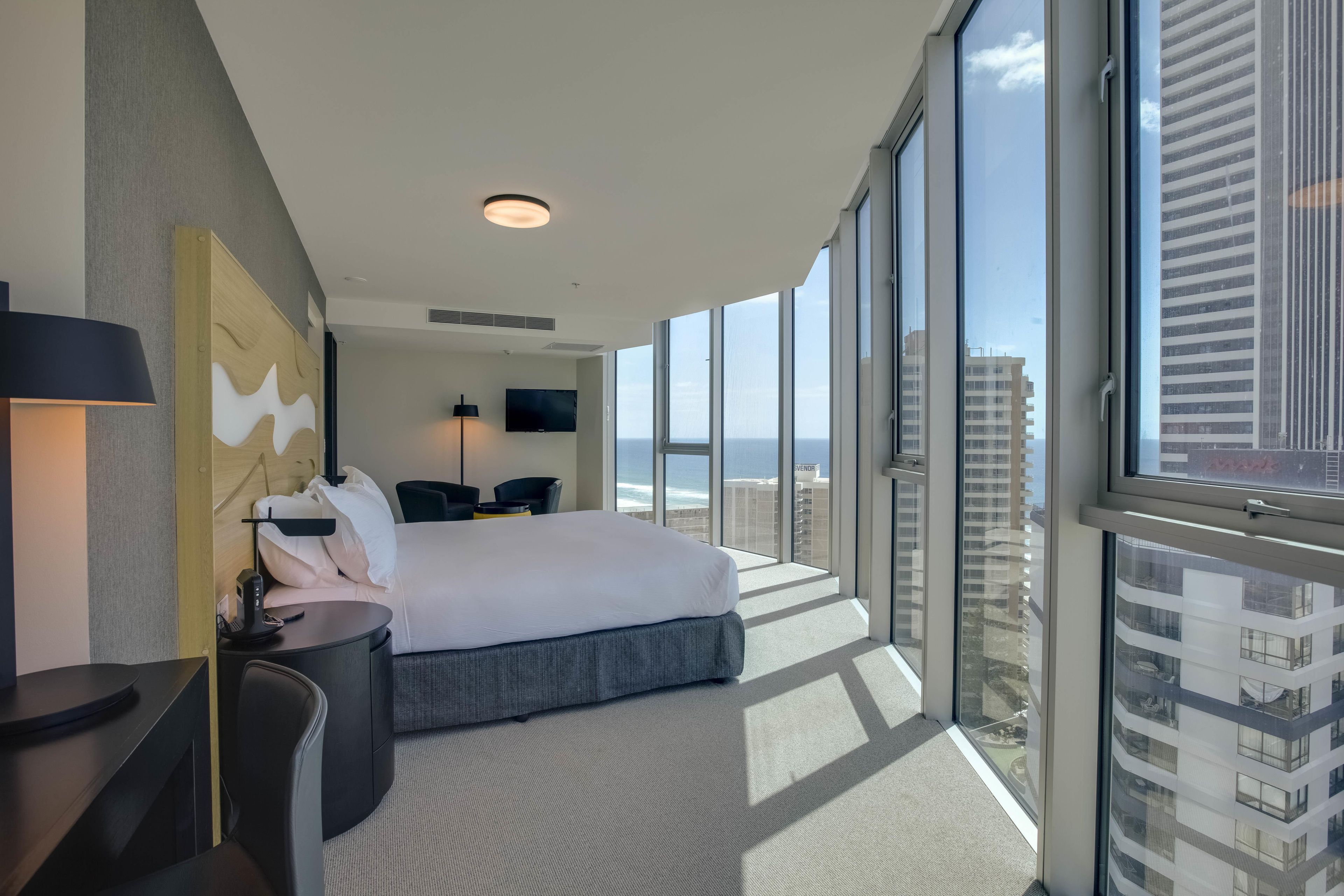 Una habitación de hotel moderna con una cama grande, un escritorio, una silla, un televisor de pantalla plana y vistas de la ciudad.