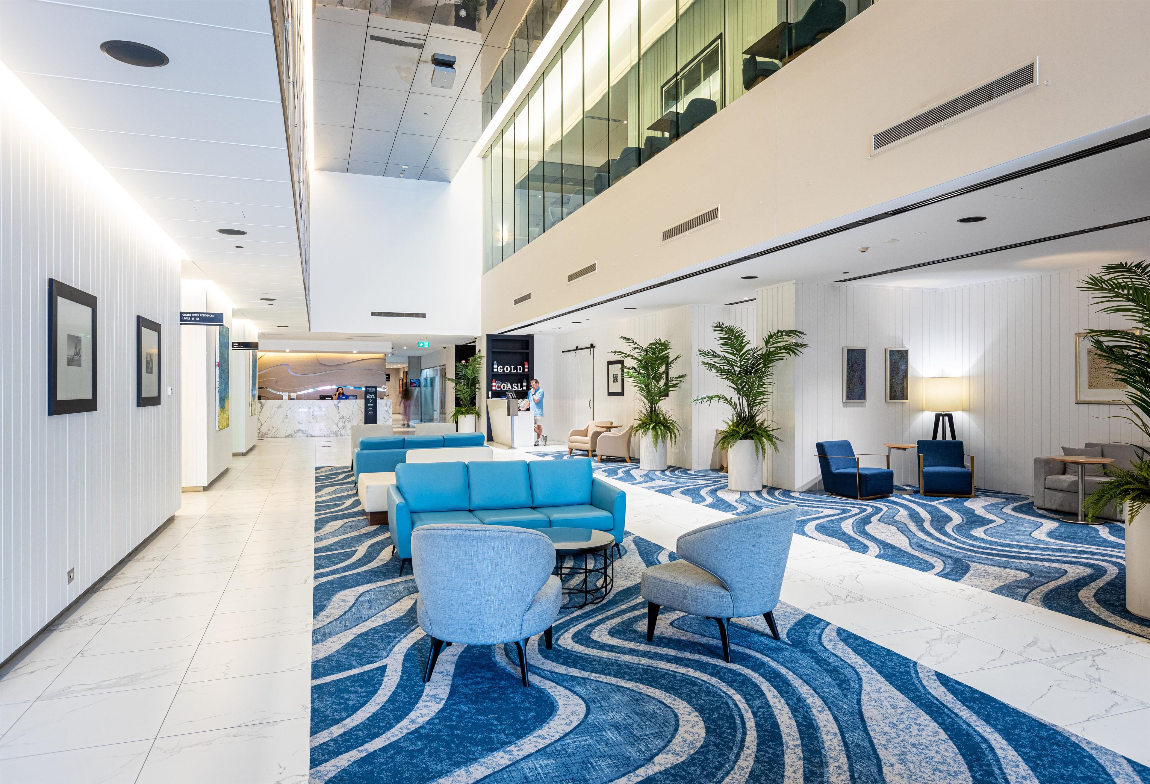 A modern hotel lobby with a reception desk, seating area, and a large blue patterned carpet.