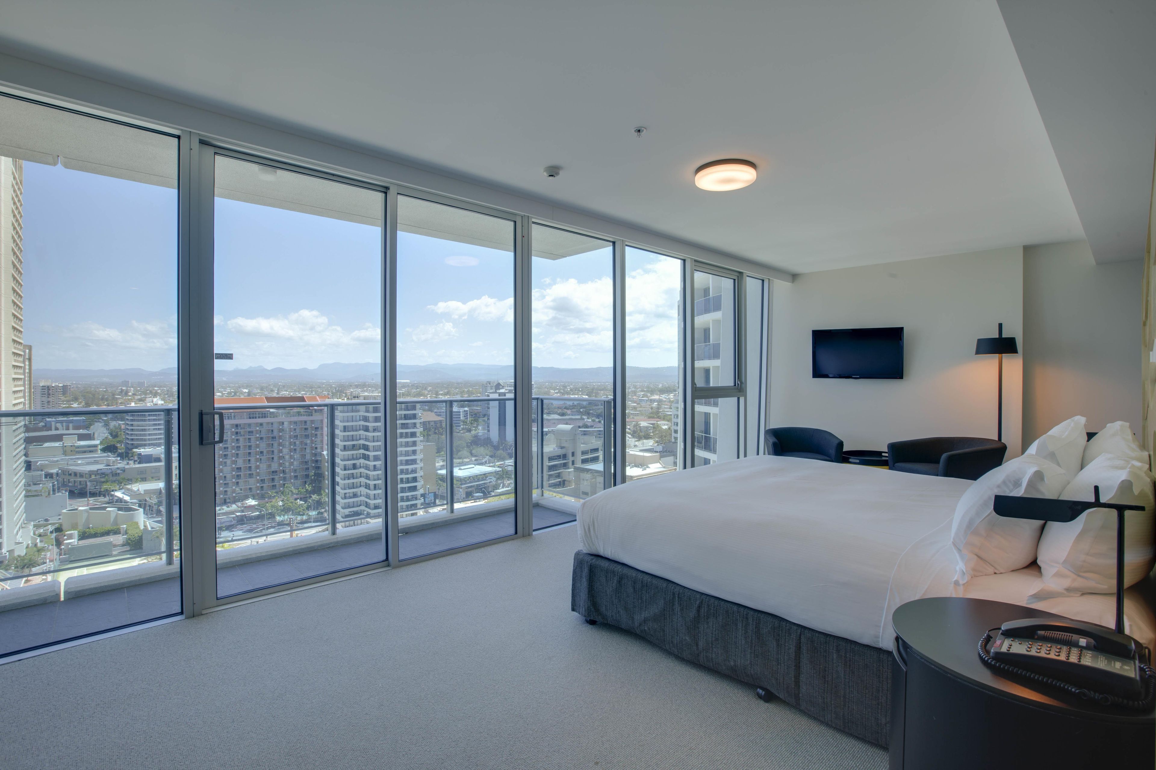 Una habitación de hotel moderna con una cama grande, un televisor de pantalla plana, un escritorio y vistas de la ciudad a través de ventanales de suelo a techo.