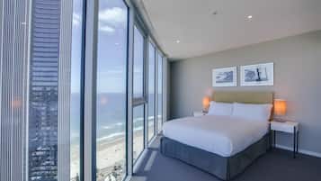 Une chambre d’hôtel dotée d’une grande fenêtre donnant sur une plage et un immeuble imposant. On y trouve un lit avec du linge de lit blanc et deux tables de chevet avec des lampes.