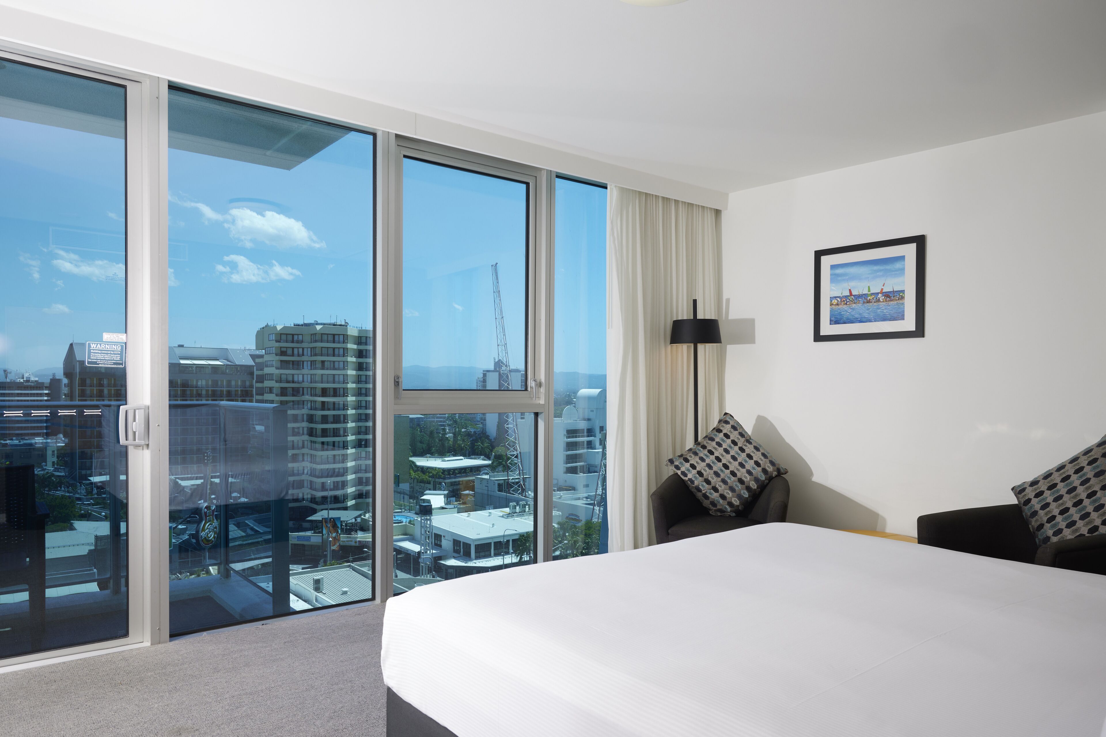 A hotel room with a large bed, a desk, and a chair. There is a large window with a view of the city.