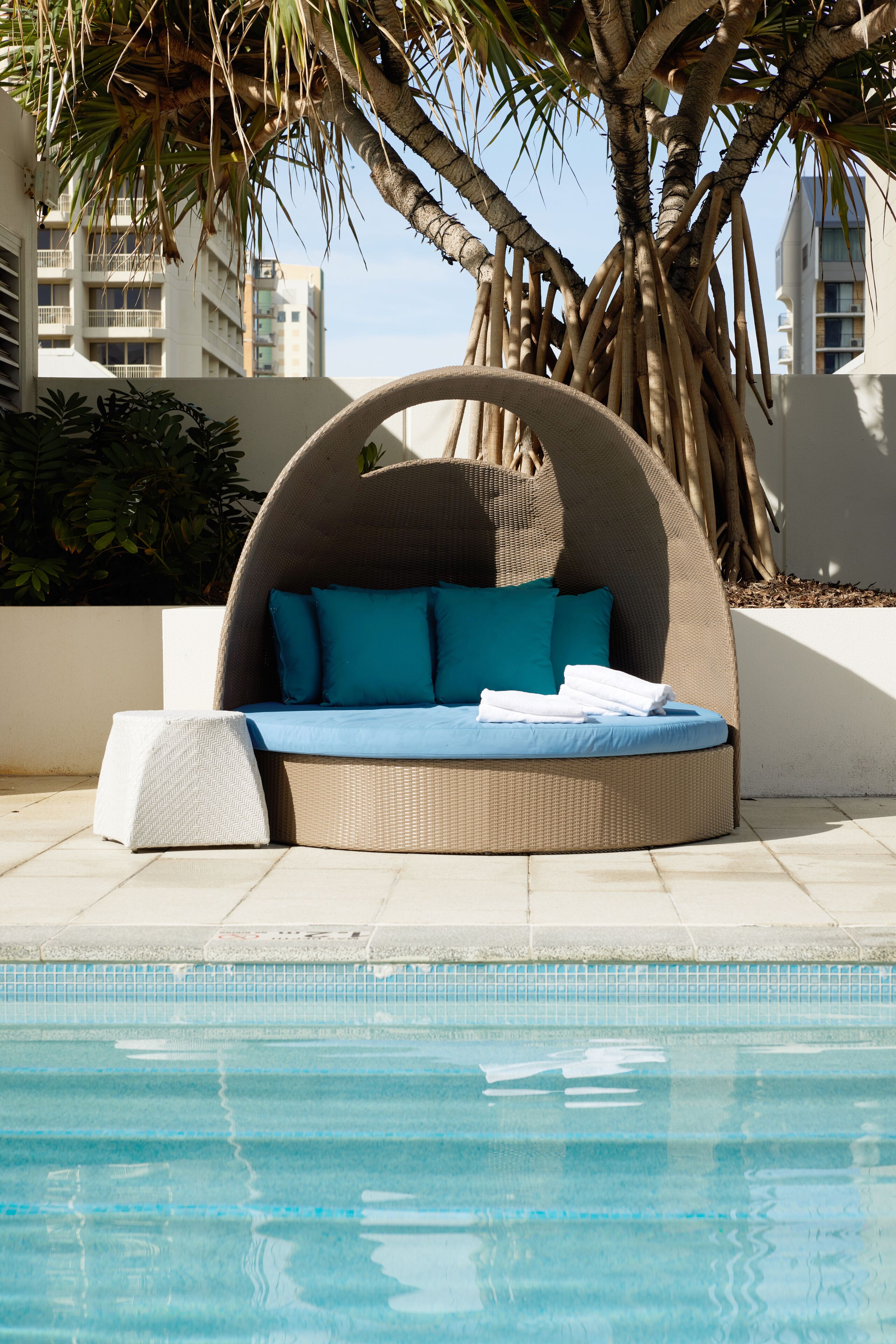 Uma área de lounge à beira da piscina com um sofá circular de vime, um banquinho branco e uma piscina com águas azuis cristalinas.