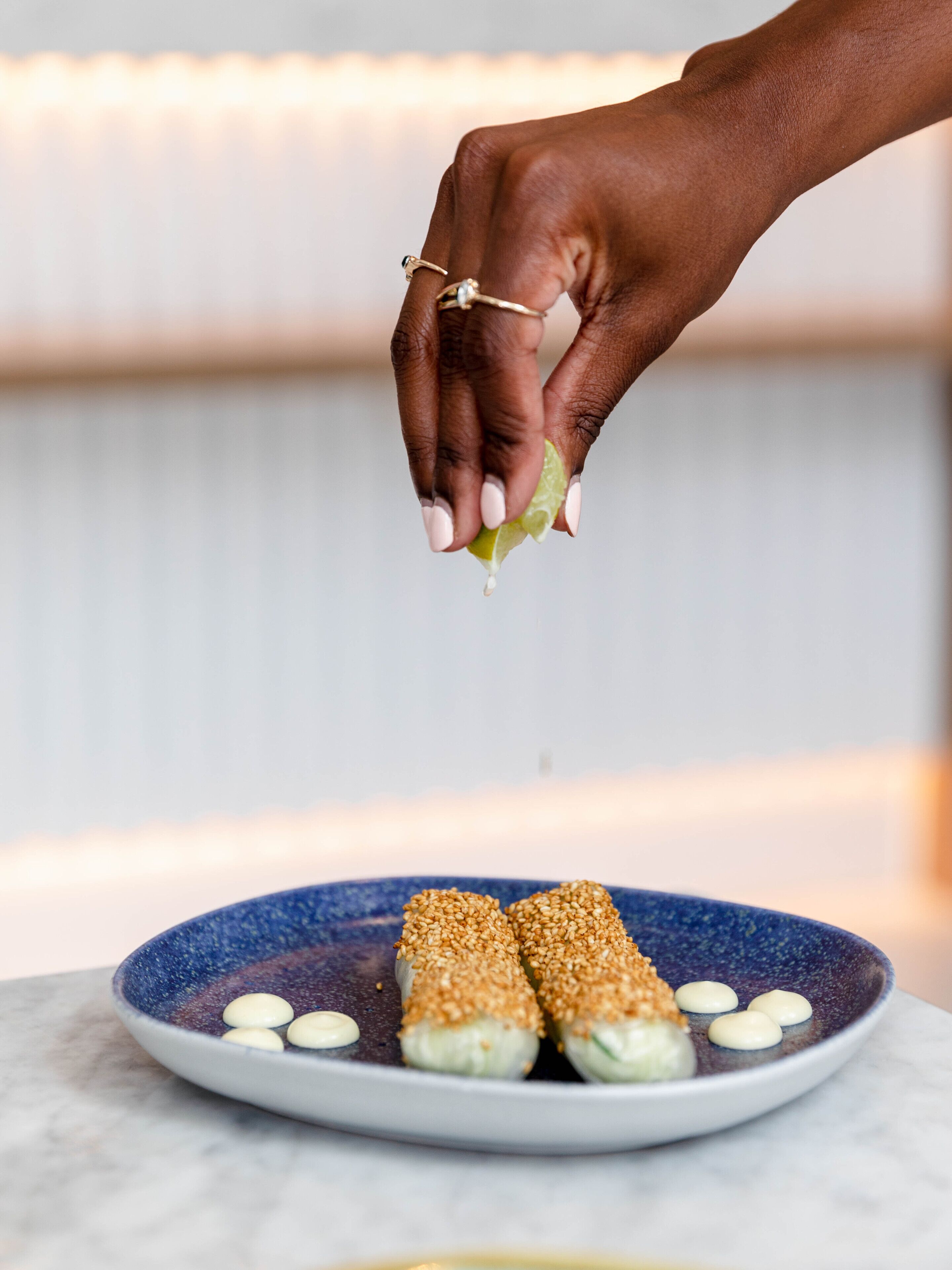 A hand sprinkling a green condiment onto a plate of food with sesame seeds and a white sauce.