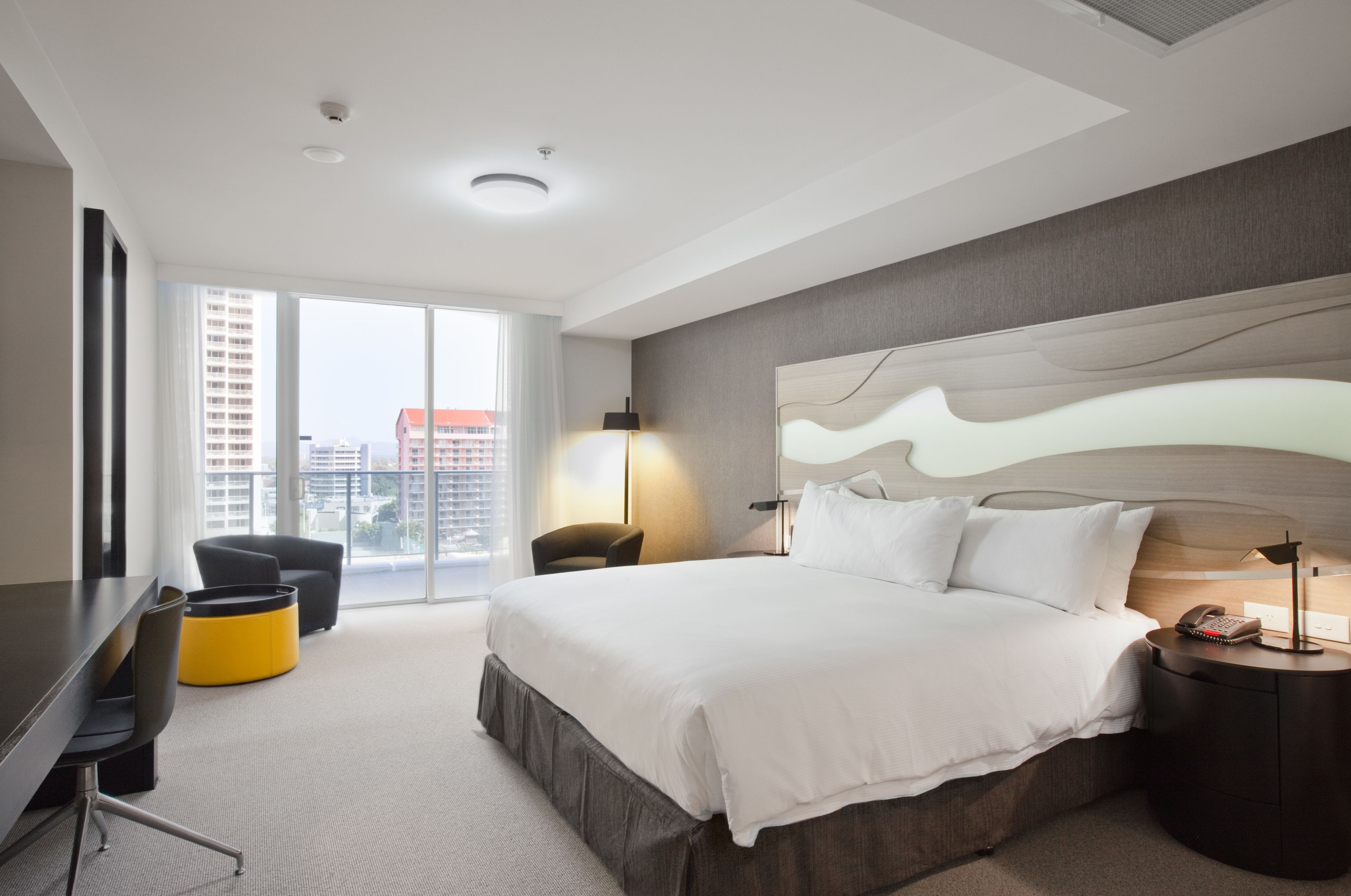Ein modernes Hotelzimmer mit einem großen Bett, einem Schreibtisch, einem Sessel und Blick auf die Stadt durch große Fenster.