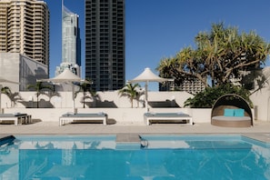 Una piscina con sillas de descanso y sombrillas, rodeada de palmeras y edificios modernos.