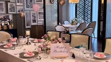 Un comedor con un cartel que dice 'Happy Birthday', mesas dispuestas con servilletas rosas y centros de mesa florales, y una pared decorada con cuadros.