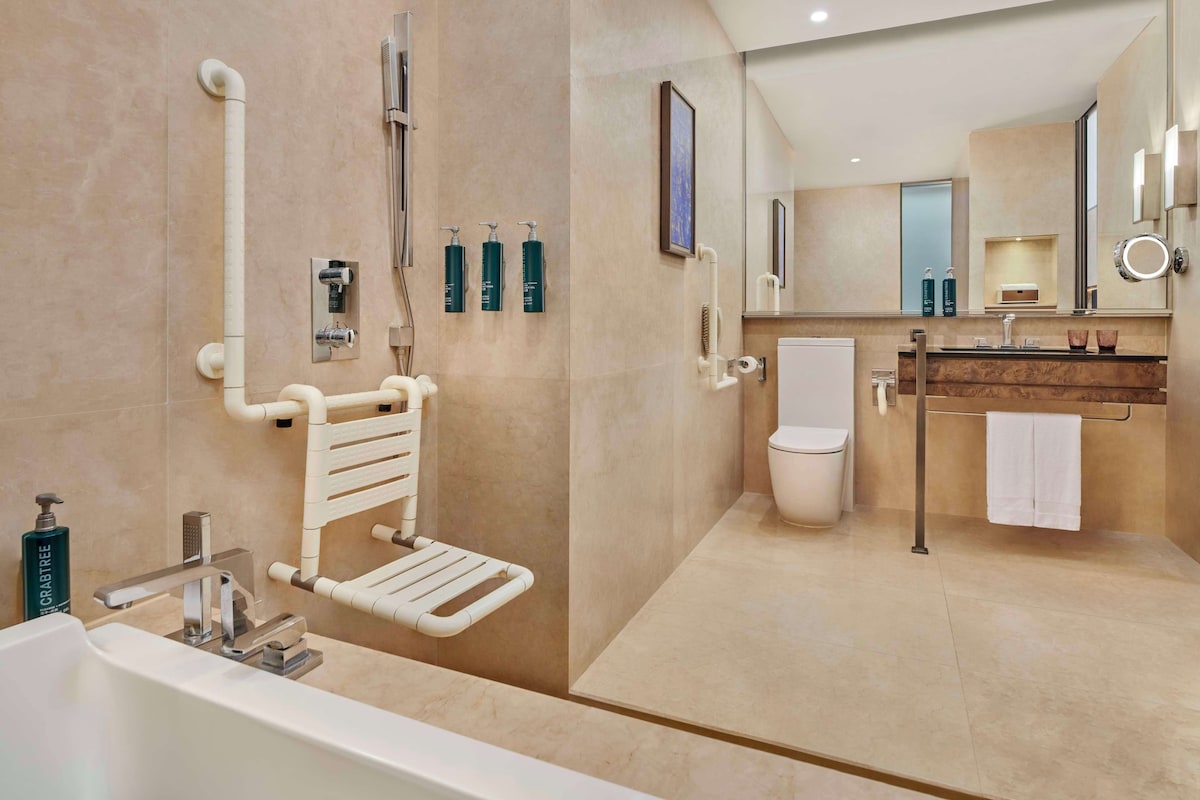 Un baño moderno con bañera, inodoro y lavamanos. Se observan barras de apoyo, un perchero para toallas y un espejo.