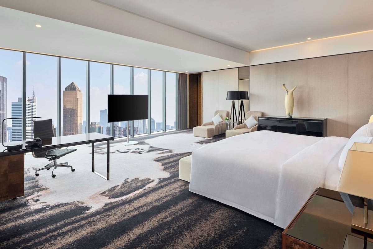 A modern hotel room with a large bed, a desk, a chair, a TV, and a city view.