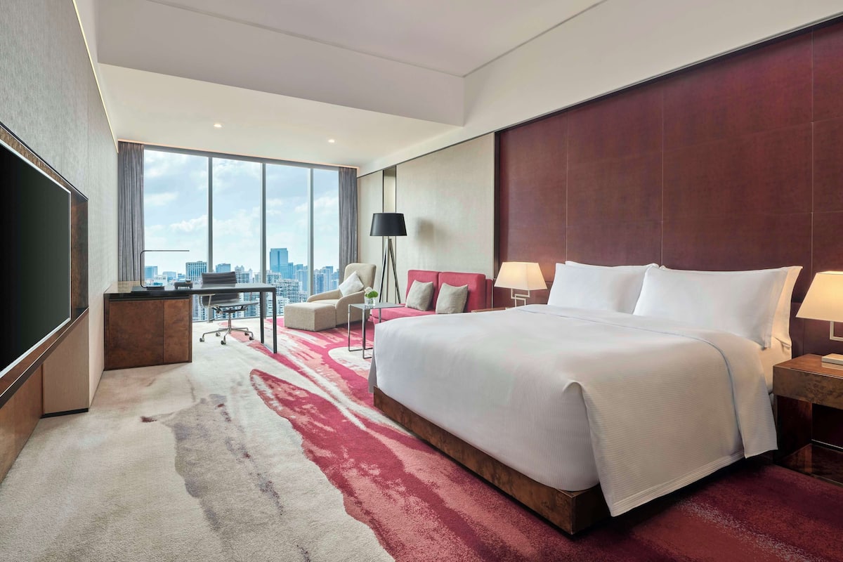 A modern hotel room with a large bed, a desk, a chair, and a view of the city skyline through large windows.