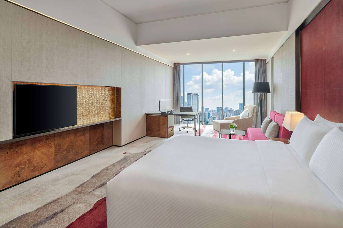 A modern hotel room with a large bed, a flat-screen TV, a desk with a chair, and a view of the city skyline through large windows.