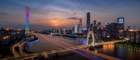 Une vue urbaine au crépuscule, avec des bâtiments illuminés, une rivière et un pont dont les lumières tracent des lignes dans le ciel.