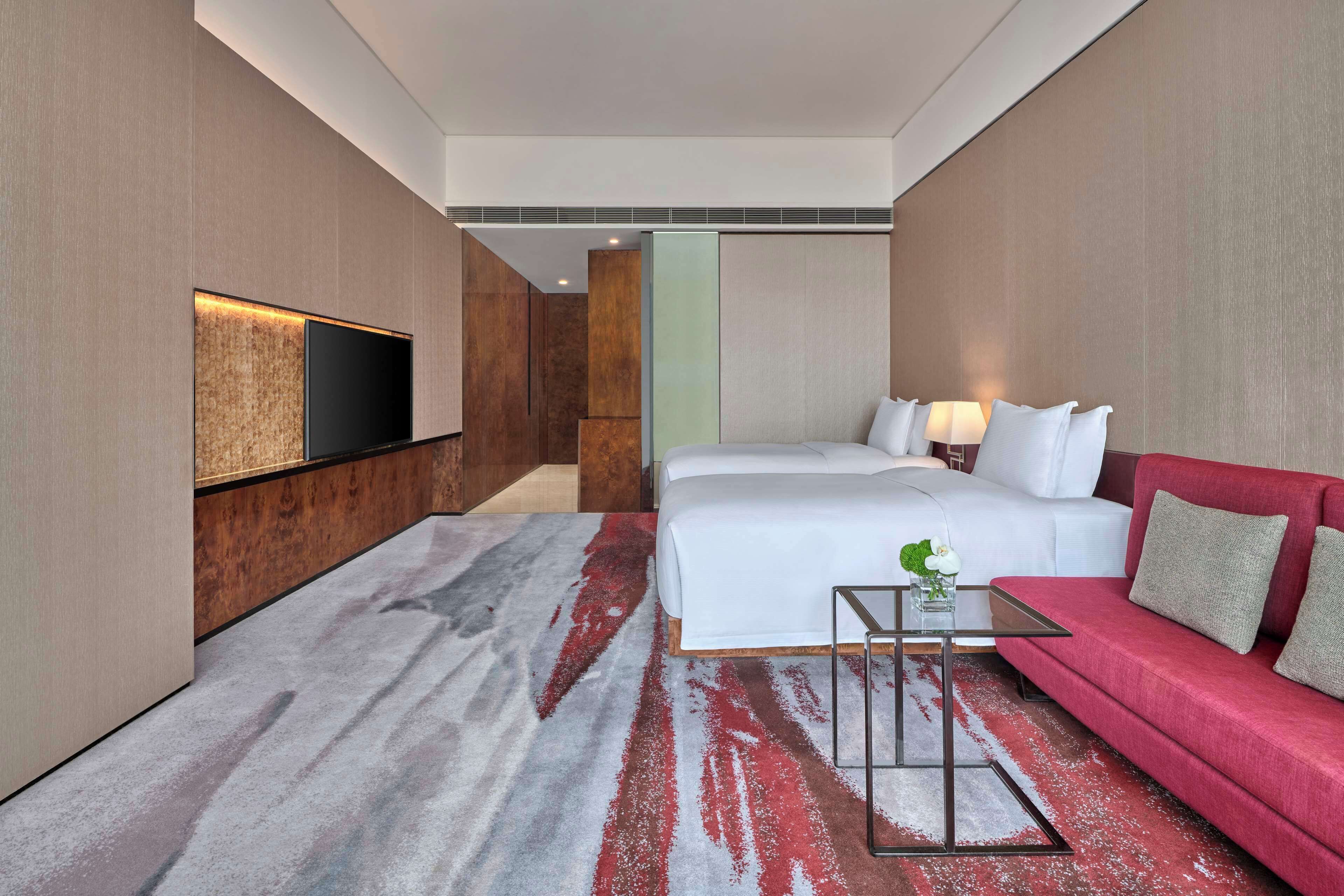 A hotel room with a bed, a red sofa, a TV mounted on the wall, and a small table with a vase.