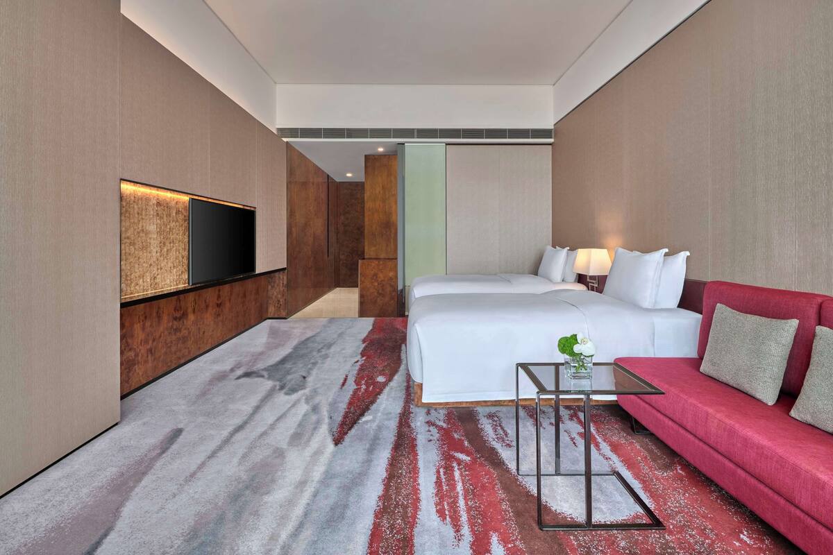 Habitación de hotel con cama, sofá rojo, televisor montado en la pared y una mesita con un jarrón.